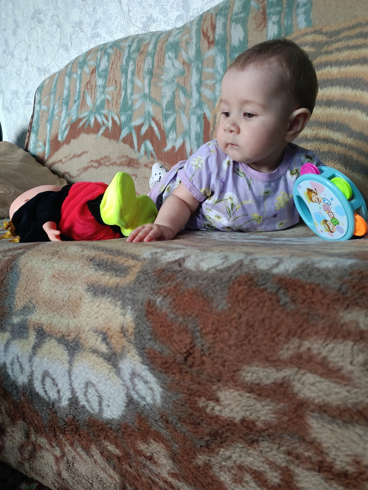 In Kamennoye, Russia, a baby lies on a couch with a doll and a toy. The baby, facing right, wears a lavender shirt with a floral pattern. A doll, dressed in red and black with yellow accents, is positioned to the baby's left. A blue tambourine toy, featuring colorful images, sits to the baby's right. The couch is covered with a patterned blanket.