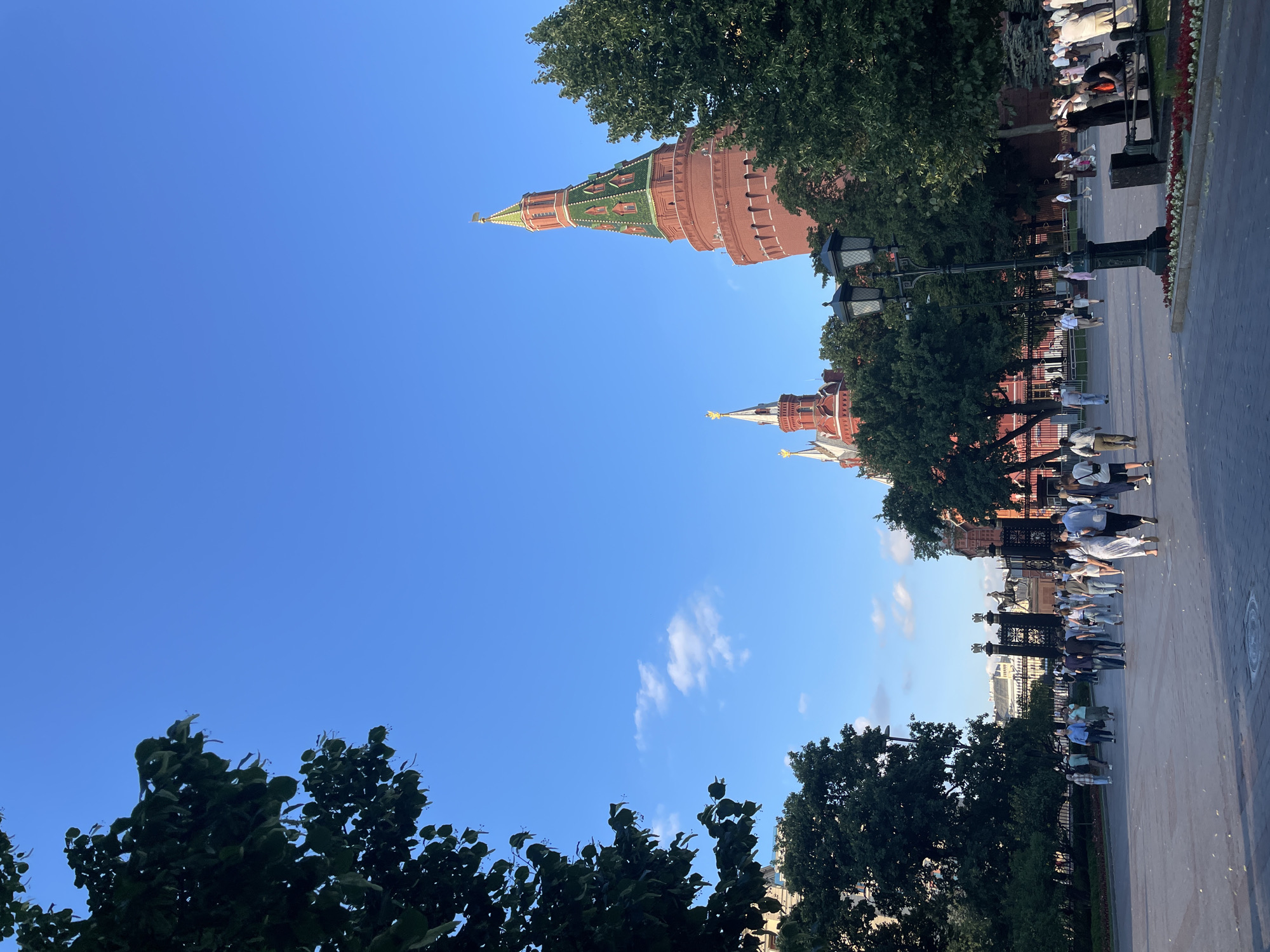 This image depicts a clear blue sky over a cityscape with several tall, red brick towers topped with green roofs and golden spires. Trees with lush green foliage are interspersed throughout the scene, some in the foreground, others framing the towers. In the middle ground, a group of people are walking along a paved walkway. Some are dressed in light-colored clothing, suggesting a warm day. Lampposts are visible along the walkway. The scene appears to be a public square or park area. The time of day is likely daytime, given the bright sky. The weather seems pleasant and sunny, with a few wispy clouds. The architectural style of the towers suggests a historical or governmental location, consistent with Moscow, Russia. The people are casually dressed and appear to be tourists or locals enjoying a walk. No visible text is present in the image.