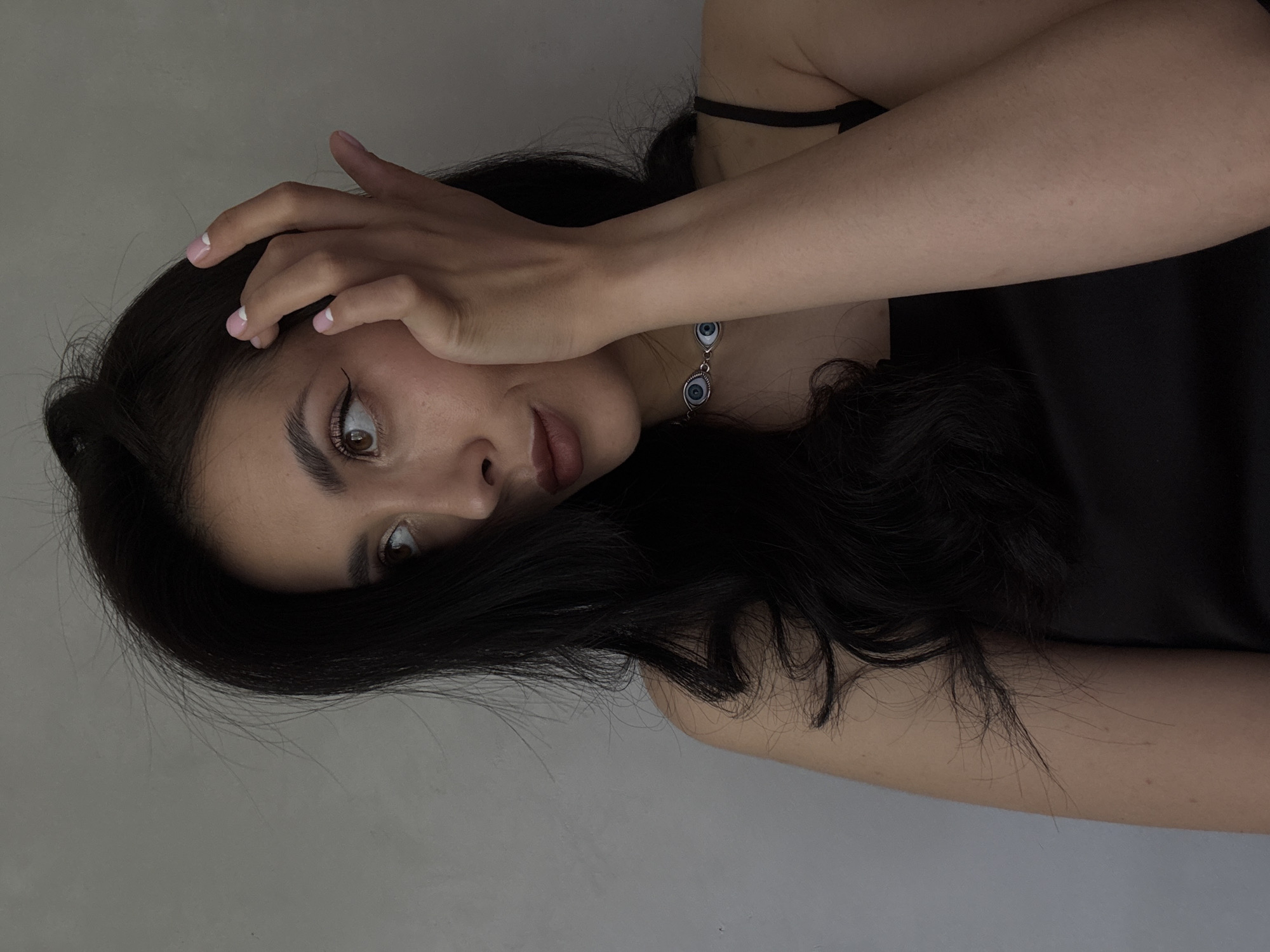 The image features a young woman with long, dark, wavy hair, captured in an indoor setting. She is lying down, positioned with her head towards the left side of the frame, and her body extending to the right, looking towards the viewer. Her right hand is raised, partially covering her forehead and the top of her face, with her fingers slightly splayed.
Her eyes are wide open, a light brown color, conveying a sense of surprise, alarm, or possibly playful shyness. She has dark, well-defined eyebrows and visible black eyeliner. Her lips are slightly parted. She is wearing a black top or dress with thin shoulder straps. Around her neck, a silver-toned necklace is visible, adorned with two distinct evil eye charms, each featuring a prominent blue iris and a black pupil. Her fingernails are manicured with a pink base and white tips.
The background is a plain, light grey wall, suggesting a simple, uncluttered indoor environment. The lighting is somewhat dim, creating soft shadows that add depth to the image. There is no visible text in the media. The location context provided is Votkinsk, Russia.