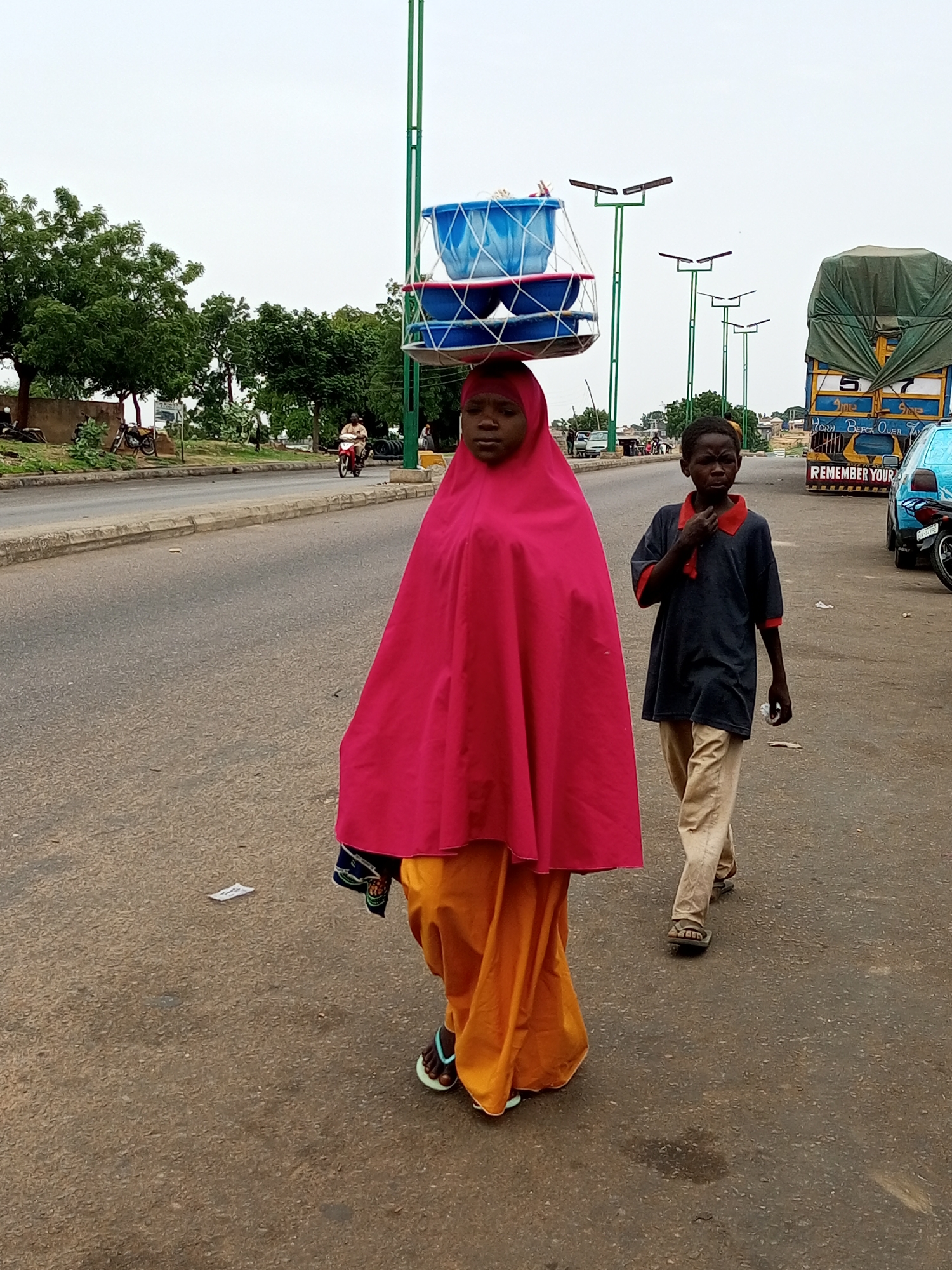 The image depicts a street scene in Gombe, Nigeria, focusing on two individuals walking along a paved road. In the foreground, a young girl is seen from the chest up, her head adorned with a stack of blue plastic basins secured by a metal frame. She is wearing a bright pink head covering and an orange skirt. To her right and slightly behind, a young boy walks alongside, wearing a dark blue shirt with red trim and light brown trousers. He is looking towards the camera with his hand near his chin. The road they are walking on is asphalt, with a concrete curb on the left. In the background, trees line the side of the road, and several streetlights with solar panels are visible. A large truck with a green tarp covering its load is also present, as is a blue car. The sky is overcast.