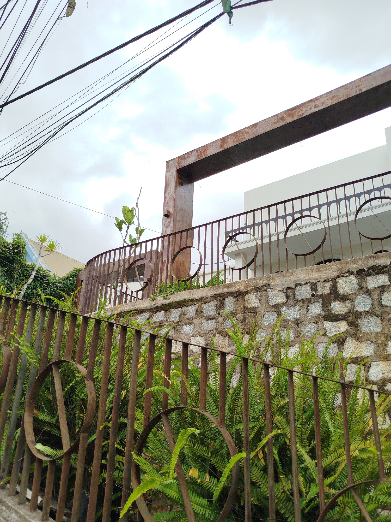 The image captures an overcast sky over a property in Da Lat, Vietnam. The foreground features a rusty metal fence with circular accents, intertwined with lush green ferns. Behind the fence, a stone wall supports a raised patio or balcony area. This area is bordered by a decorative metal railing with repeating arched patterns and circular openings. Beyond the railing, a modern white building is visible, with a large, rusty metal beam structure extending overhead. Overhead power lines are strung across the upper portion of the image.