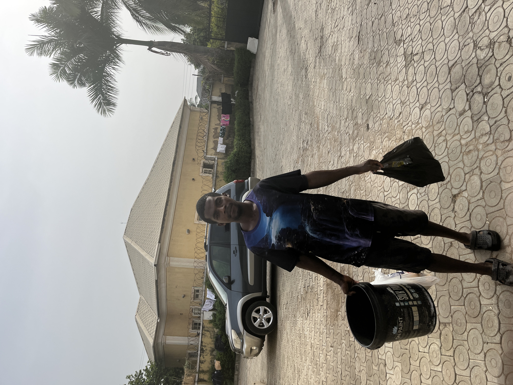 The image shows a man standing outdoors in a paved residential area, looking directly at the camera with a neutral expression. He has dark skin and is wearing a dark t-shirt with a blue and purple graphic design, black shorts, and dark clogs or sandals. There are some paint splatters visible on his clothing.
In his left hand, the man holds a black plastic bucket. Partial text on the bucket reads "SILK PU" and "ON PAINT", indicating it is likely a paint container. In his right hand, he holds a black plastic bag containing visible items.
Behind the man, a dark-colored SUV is parked. Further back, a large house with light yellow walls and a distinctive light-colored, patterned roof is prominent. The property features a fence topped with barbed or razor wire, behind which green bushes grow. Several pieces of laundry are hanging on a line or fence in the background. A tall palm tree with green fronds is visible against a bright, hazy sky in the upper left portion of the image.
The ground is paved with light-colored, interlocking tiles that feature a repetitive circular pattern. The lighting suggests it is daytime, under a hazy or overcast sky, common in many tropical climates. The scene depicts a typical residential compound.