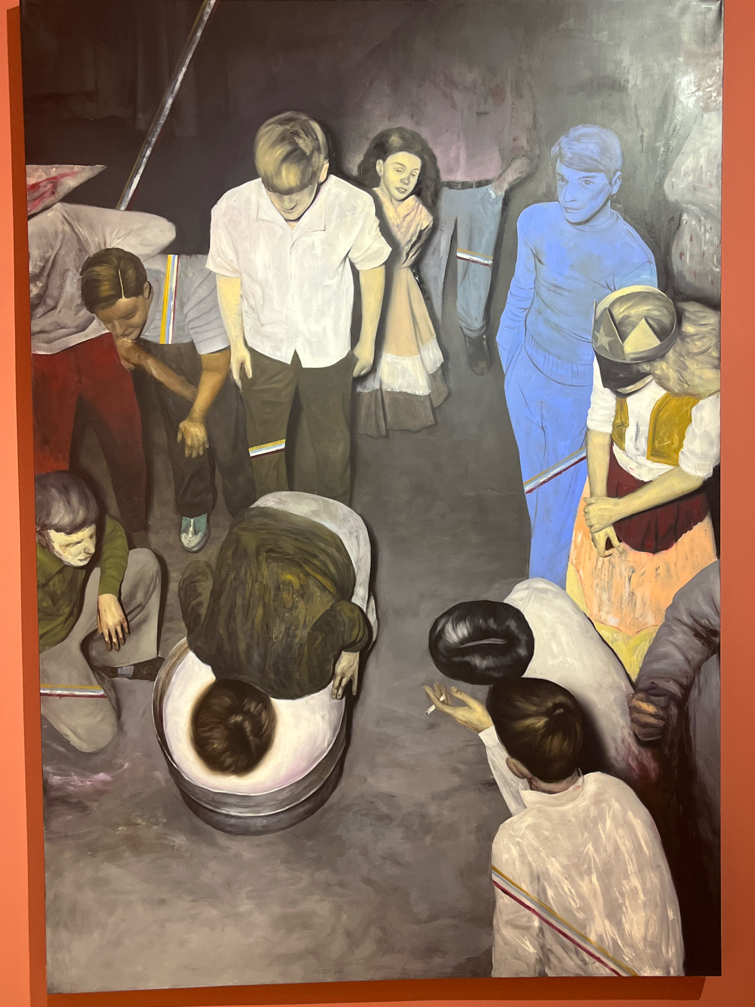 The painting depicts a group of individuals, appearing to be children and adolescents, in a dimly lit, enclosed space. The figures are rendered in a semi-realistic style with muted tones, though some figures, like the one on the right, are painted in a distinct blue. Several individuals are depicted in various poses: one is bent over a large, metallic basin, with their head submerged in what appears to be a white liquid; another is crouched low, seemingly observing the person in the basin; a figure in the center wears a white shirt and looks downwards; and a girl in a light-colored dress stands to the side.
In the foreground, a person with dark hair is shown from behind, wearing a white shirt and holding a lit cigarette. Other figures are visible in the background, their features less distinct. The environment appears to be an indoor space with a dark, textured background, possibly suggesting a stage or an industrial setting. The overall atmosphere is somber and possibly unsettling, with a focus on the interactions and postures of the young people.
This artwork is displayed on an orange wall, indicating it is likely part of an exhibition. This specific location is within the **National Museum of Montenegro in Podgorica, Montenegro**, specifically in the section dedicated to contemporary art.