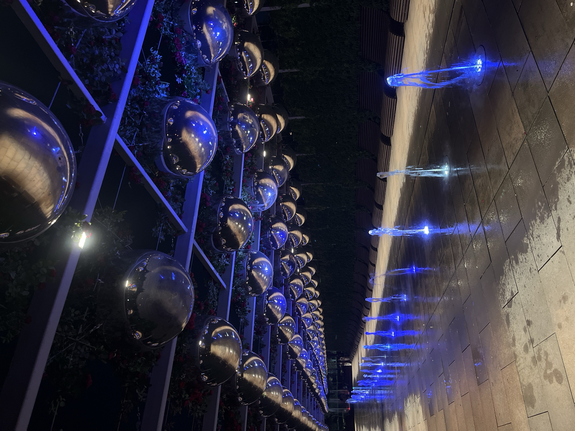 The image depicts an outdoor promenade at night, illuminated by blue and white lights. A long row of reflective, spherical objects hangs from a pergola-like structure adorned with foliage and red flowers. Below, a series of water fountains spray upwards, casting blue and white light onto the wet pavement. In the background, rows of dark wooden benches are visible, backed by a dense, dark green hedge. The scene appears to be located in Krasnodar, Russia, likely in a park or public square. No people are clearly visible, though a few figures are faintly discernible in the far distance on the left. The overall atmosphere is serene and visually striking, emphasizing the interplay of light, water, and reflective surfaces.