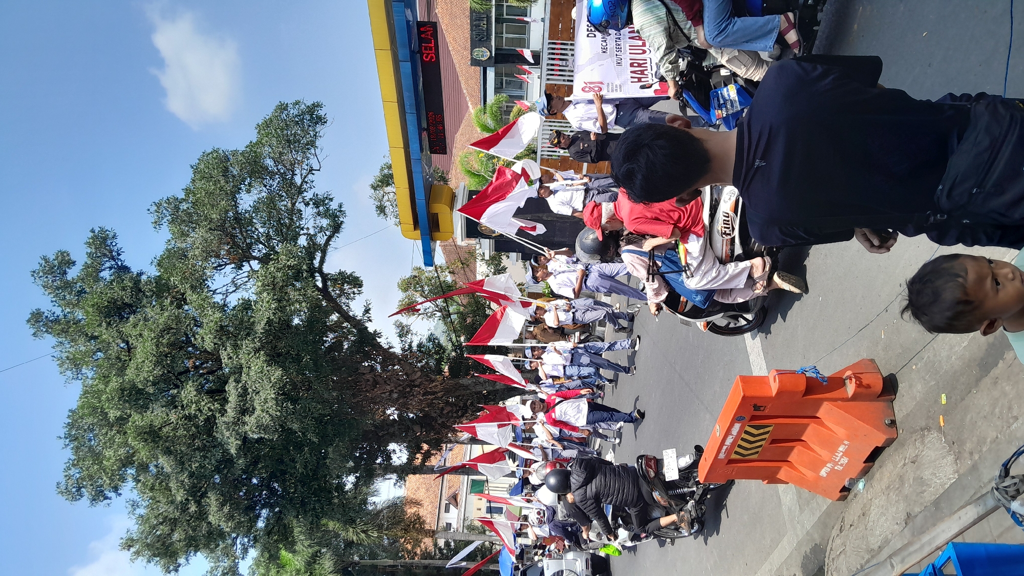 The image depicts a parade in the city of Sumedang, Indonesia, likely celebrating a national holiday. A procession of students, many wearing school uniforms and carrying Indonesian flags, marches down a street. Some students are riding motorcycles, while others walk alongside. In the background, a building with a yellow awning and the word "SELAMAT" is visible, possibly a government office or public facility. The scene is set under a bright blue sky with scattered clouds, and a large, leafy tree dominates the left side of the frame. Orange traffic barriers are placed along the side of the road, suggesting crowd control measures. People are observing the parade from the sidewalk, with a few individuals in the foreground facing away from the camera.