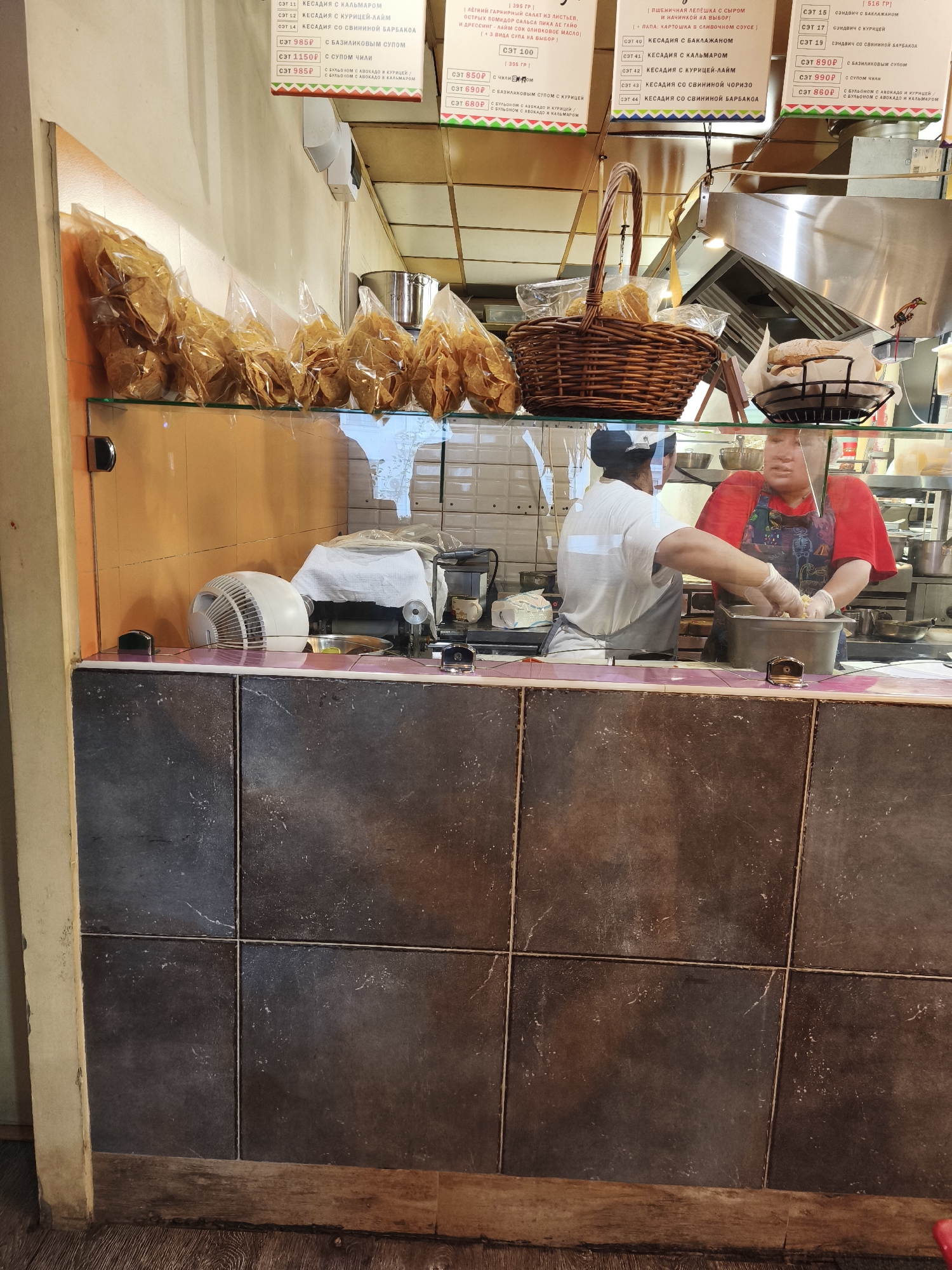 The image depicts the interior of a food establishment in Vladivostok, Russia, specifically a casual eatery or takeaway counter. In the foreground, the dark tiled counter serves as a base for a glass display case. Behind the glass, several plastic bags filled with what appear to be tortilla chips or similar fried snacks are neatly arranged on an orange-colored shelf. A wicker basket containing bread items sits on the same shelf.
In the mid-ground, two individuals are visible behind a transparent protective barrier. One person, wearing a white t-shirt and an apron, is partially obscured and appears to be preparing food. The other individual, wearing a red t-shirt and a patterned apron, is actively engaged in handling food items within a stainless steel container. Both individuals are wearing clear plastic gloves. Various kitchen appliances and containers are visible in the background, including stainless steel pots and what seems to be a food preparation station. The overall environment suggests a functional, operational food service setting.