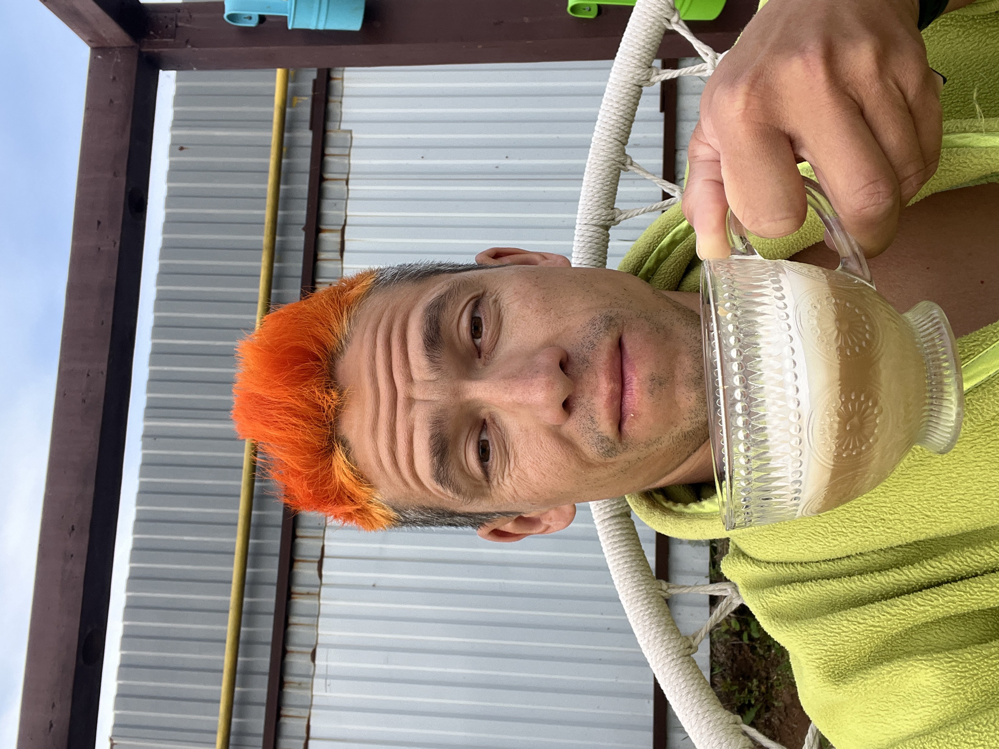 A person with orange hair is sitting in a white hammock, holding a glass mug filled with a light-colored liquid. The person is wearing a lime green fleece jacket. Behind them is a corrugated metal fence, a brown wooden frame, and a yellow pipe. Above the frame, there are small, colorful containers. The photo was taken in Chaikovsky, Russia.