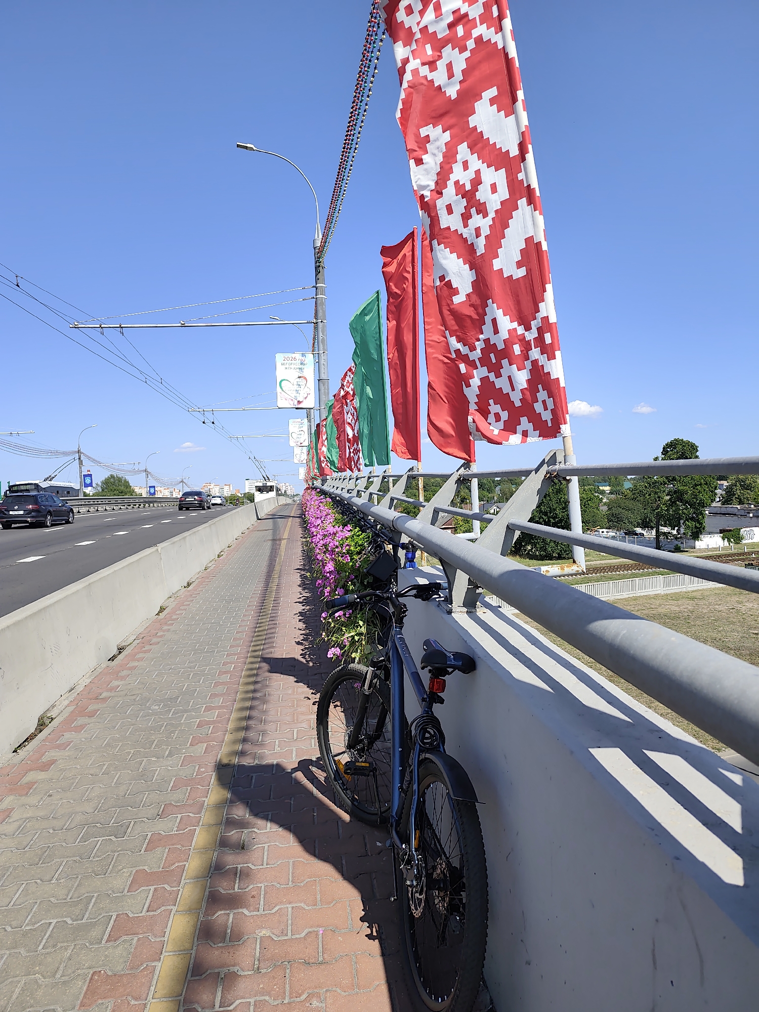 On a clear, sunny day, a blue bicycle is parked on a pedestrian walkway beside a highway. The walkway is paved with red and grey interlocking bricks and bordered by a concrete barrier and a metal railing. Pink flowers are planted along the base of the barrier. Large red and white patterned flags are strung along poles above the walkway, interspersed with plain green and red flags. Overhead, trolleybus wires stretch across the scene. In the background, cars travel on the highway, and beyond that, buildings and trees are visible. A signpost with informational posters is also present. The image appears to be taken from the pedestrian area of a bridge, possibly in Brest, Belarus, given the national symbolism of the flags and the presence of trolleybuses.