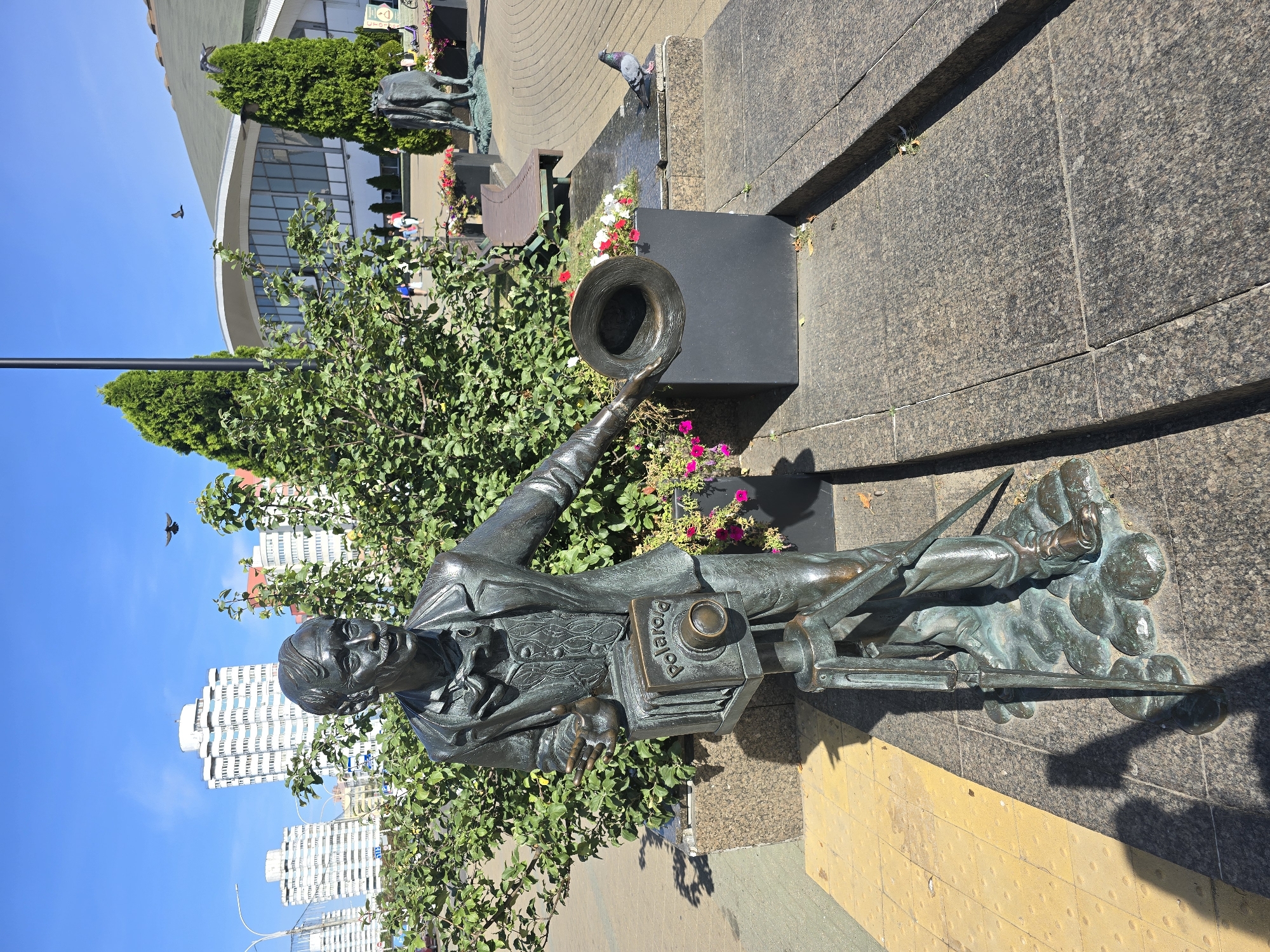 This is a full-shot, eye-level, outdoor, natural light photograph of a bronze sculpture of a man holding a Polaroid camera and a hat. The sculpture is positioned in front of a dense green bush. The man has a mustache, is wearing a suit with a waistcoat and a bow tie. The camera is mounted on a tripod, and the word "Polaroid" is visible on its casing. The sculpture's base is made of granite. To the right of the sculpture, a dark planter box with red and white flowers is visible, and beyond that, a paved area with a park bench and a statue of a cow. In the background, there are several white high-rise buildings and a large modern building with a curved roof and large windows. The sky is clear and blue, with two birds in flight. A pigeon is standing on the granite next to the planter box. This sculpture is located in Minsk, Belarus, specifically at the crossroads of Independence Avenue and Surganov Street, known as the "Minsk Square" or "Central Square."