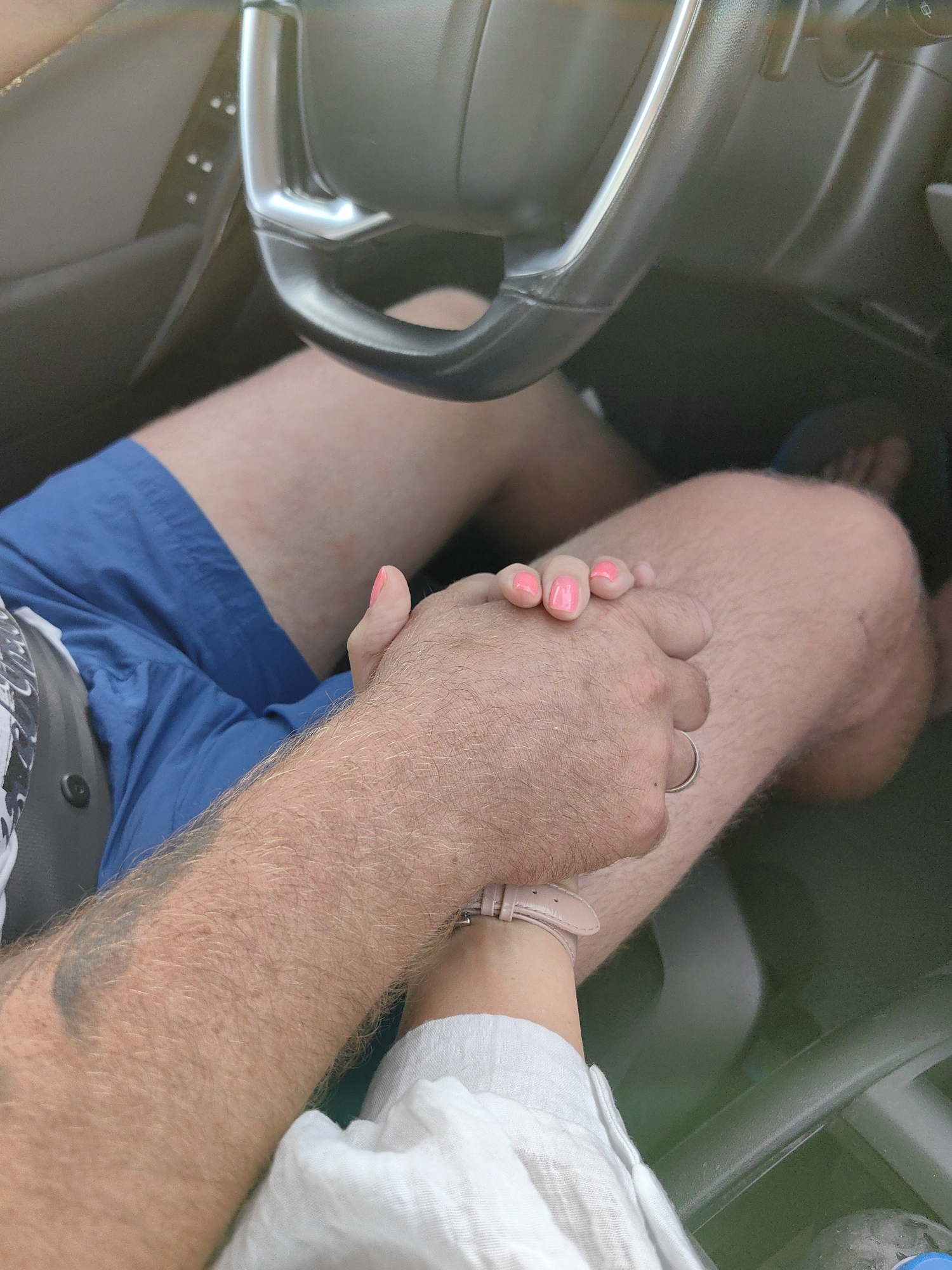 In an automobile interior, two individuals are holding hands. One person, wearing blue shorts, has their right hand on the steering wheel, while their left hand is held by the other person. The other individual's hand, adorned with bright pink nail polish, is intertwined with the first person's hairy arm. A silver ring is visible on the first person's hand, and a light-colored watch is worn on the wrist of the second person. The background shows parts of the car's dashboard and steering wheel. The setting is an unspecified street in Янушкавіцкі сельскі Савет, Belarus.