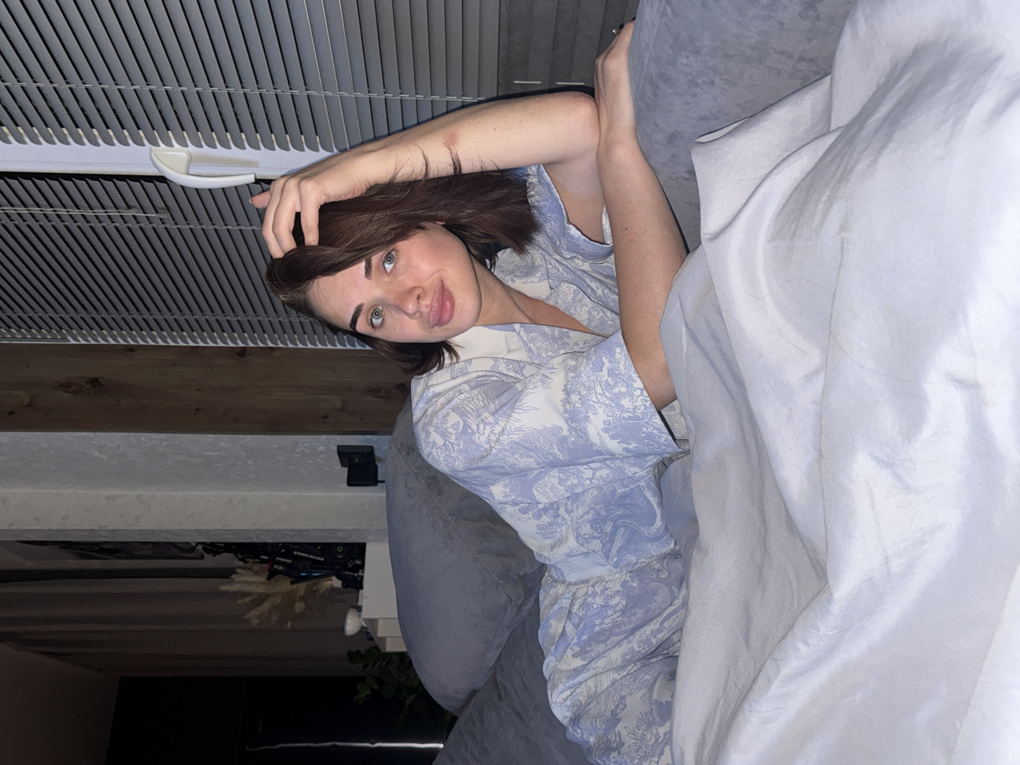 A young woman with brown hair and fair skin is lying on a gray couch, partially covered by a white blanket. She is wearing light blue patterned pajamas. She has one hand behind her head, is sticking her tongue out and looking at the camera. The background includes a window with closed blinds, and a shelf with items on it. The image appears to be taken indoors and the location is Lobanovo, Russia.