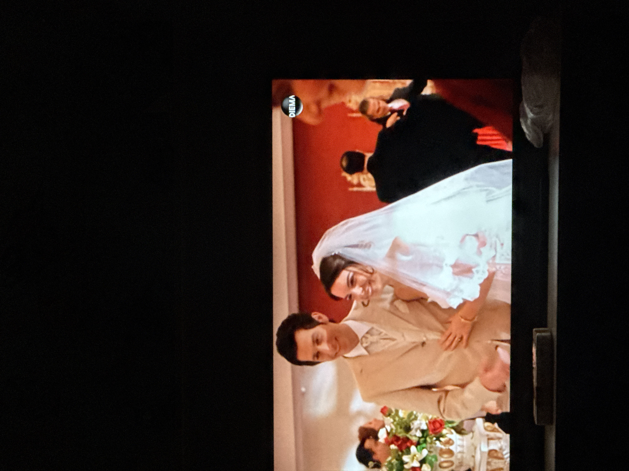 A photo of a TV screen shows a wedding scene. The bride and groom are embracing and smiling. They appear to be at a wedding reception, as there are other people and a table with flowers visible in the background. The logo "DIEMA" is in the top left corner of the TV screen, indicating the channel the image is from. The image does not contain any information about Sarnevo, Bulgaria.