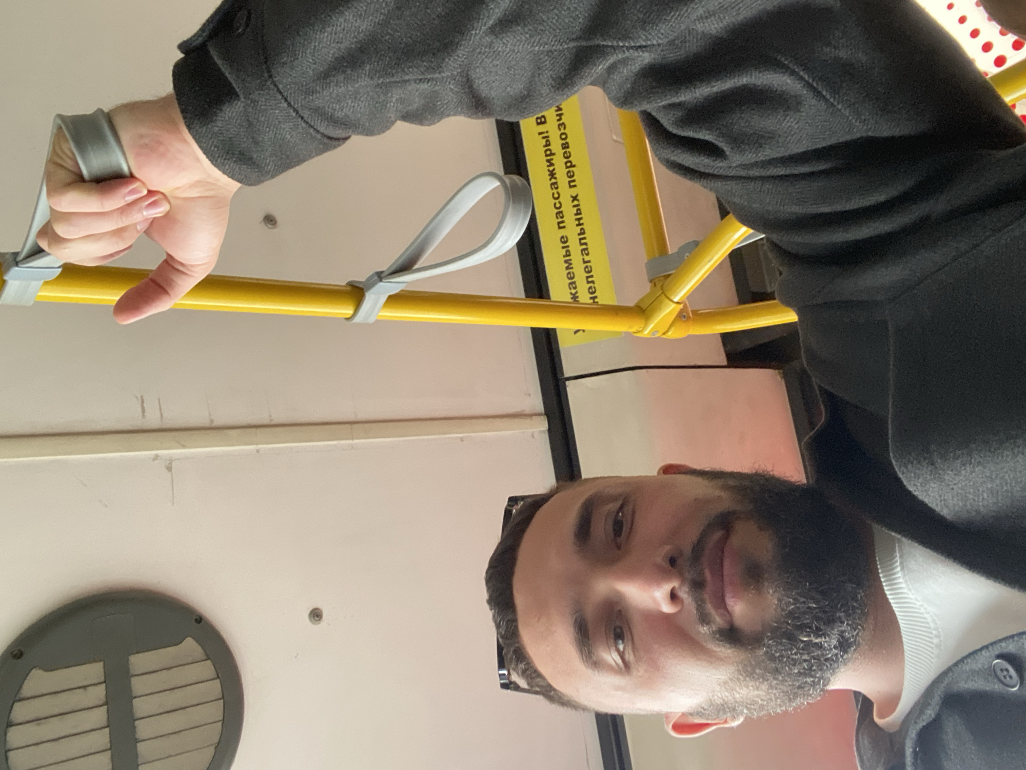 The image shows a close-up, selfie-style shot of a man with a beard, wearing a dark jacket over a white sweatshirt. He is inside a public transport vehicle, likely a bus, evidenced by the yellow pole with a grey hand strap that he is holding. In the background, a yellow sign with Russian text is visible. The man's face is the primary focus, with his left eye and eyebrow slightly obscured by sunglasses perched on his head. The environment appears to be well-lit, suggesting daytime. The location of Новое Мартьяново, Russia, is not discernible from the visual information present in the image.