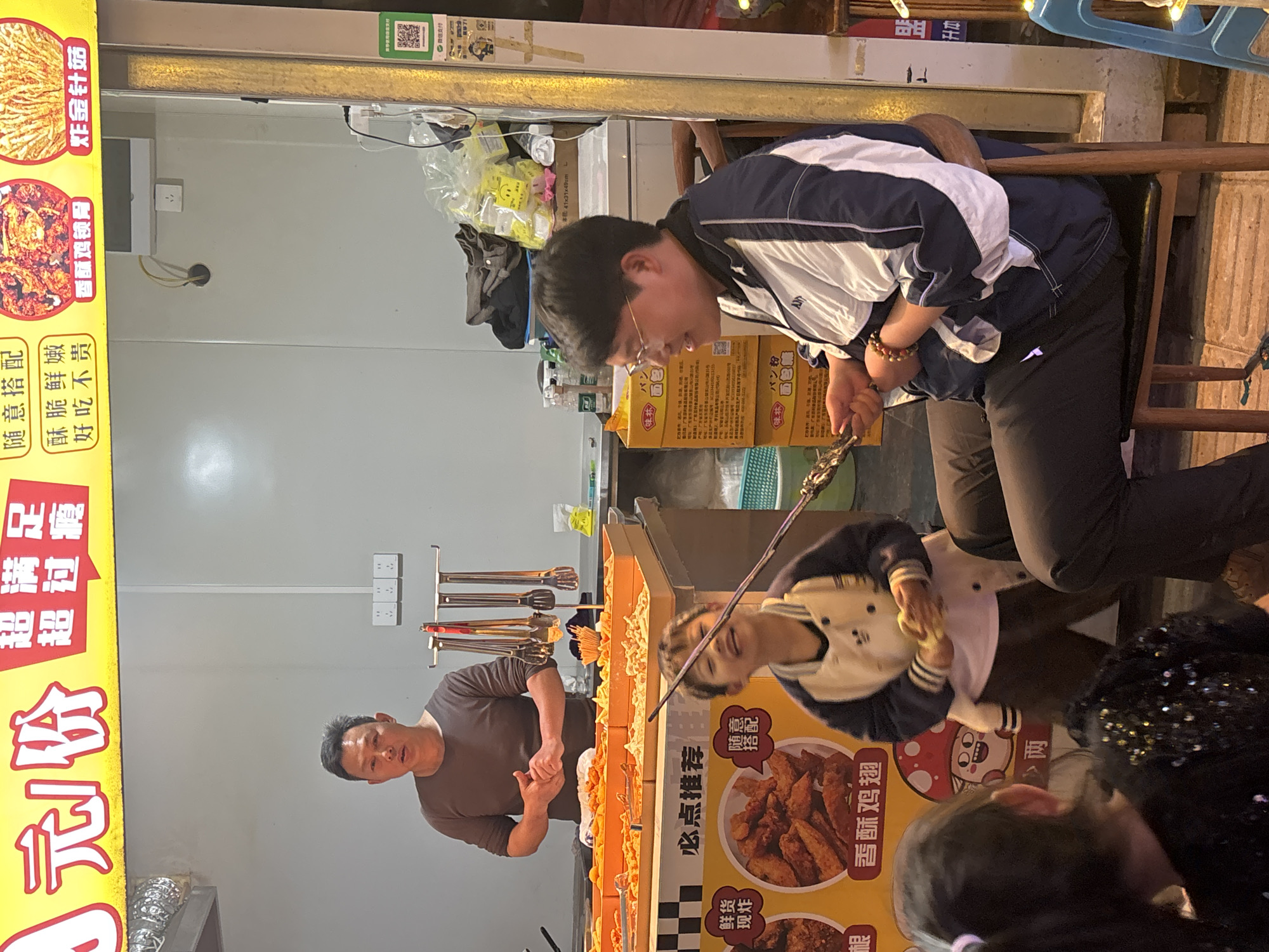 This outdoor street scene shows people gathered at a food vendor in the city of Jiangjin, China. A man is behind the counter, preparing or serving fried food. A vertical sign adjacent to him lists available items, including fried chicken wings and fried enoki mushrooms. A customer is seated on a stool and leaning forward, looking at the food. Other customers are near the counter, including a child in a white and blue jacket. The vending area is covered by a white metal shell with electrical sockets and tools. Above the food area, there are various boxes and bags.