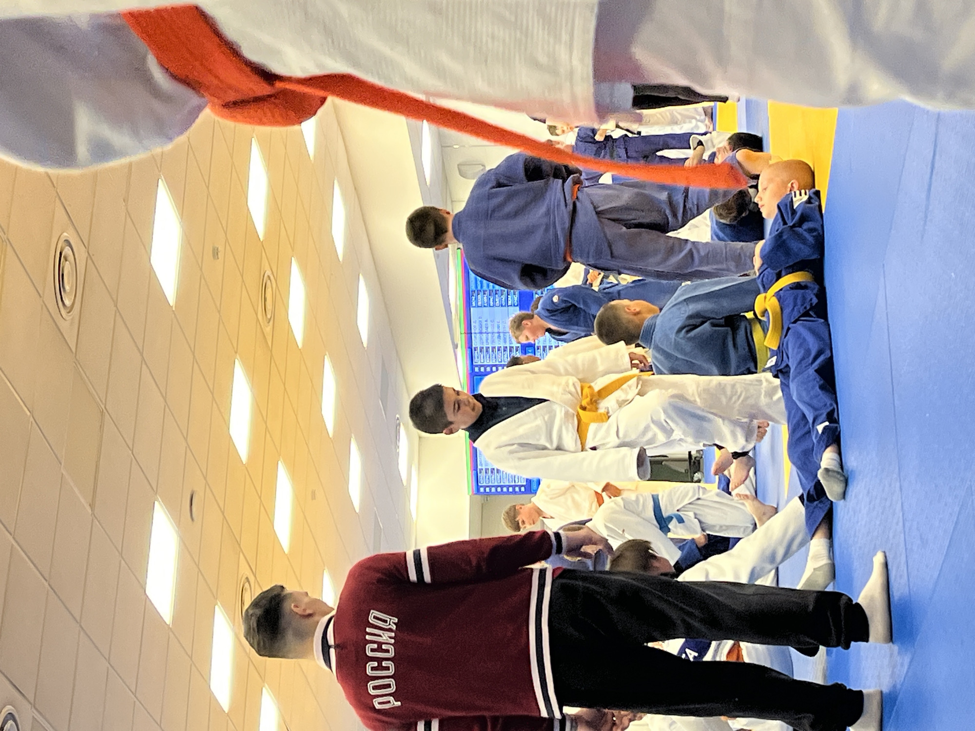 The image depicts an indoor martial arts training or event taking place in Krasnoyarsk, Russia. The scene is illuminated by numerous square, recessed LED or fluorescent lights integrated into a suspended grid ceiling of beige acoustic tiles, which also feature circular air vents. The floor is covered with blue and yellow interlocking foam mats typical of a dojo or training hall.
In the left foreground, a man with a dark, short haircut is seen from behind, wearing a maroon long-sleeved sweater with white horizontal stripes on the sleeves and the word "РОССИЯ" (Russia) emblazoned in white capital letters across the upper back. He is wearing black pants and white socks, standing with his feet apart on the blue mat.
In the central middle ground, a boy approximately 10-12 years old, dressed in a white judogi with a yellow belt, walks towards the right, looking downwards. Behind him and to his left, another young boy in a blue judogi with a yellow belt is lying on his back on the blue mat, facing towards the left side of the frame.
Further in the middle ground and background, several other individuals, mostly children and teenagers, are visible. They are predominantly dressed in either white or blue judogi, some with blue belts, engaged in various postures on the mats—some standing, some sitting or kneeling. One person in a blue judogi is seen from behind, standing erect. To the far right, a partial view of a person in a white judogi with a bright orange/red sash or scarf is visible, facing away from the camera.
In the background, a large digital display or scoreboard with columns of text, possibly names or scores, is visible against a light-colored wall, emitting blue and green light. The overall atmosphere suggests an organized sports activity, specifically related to judo or a similar martial art.