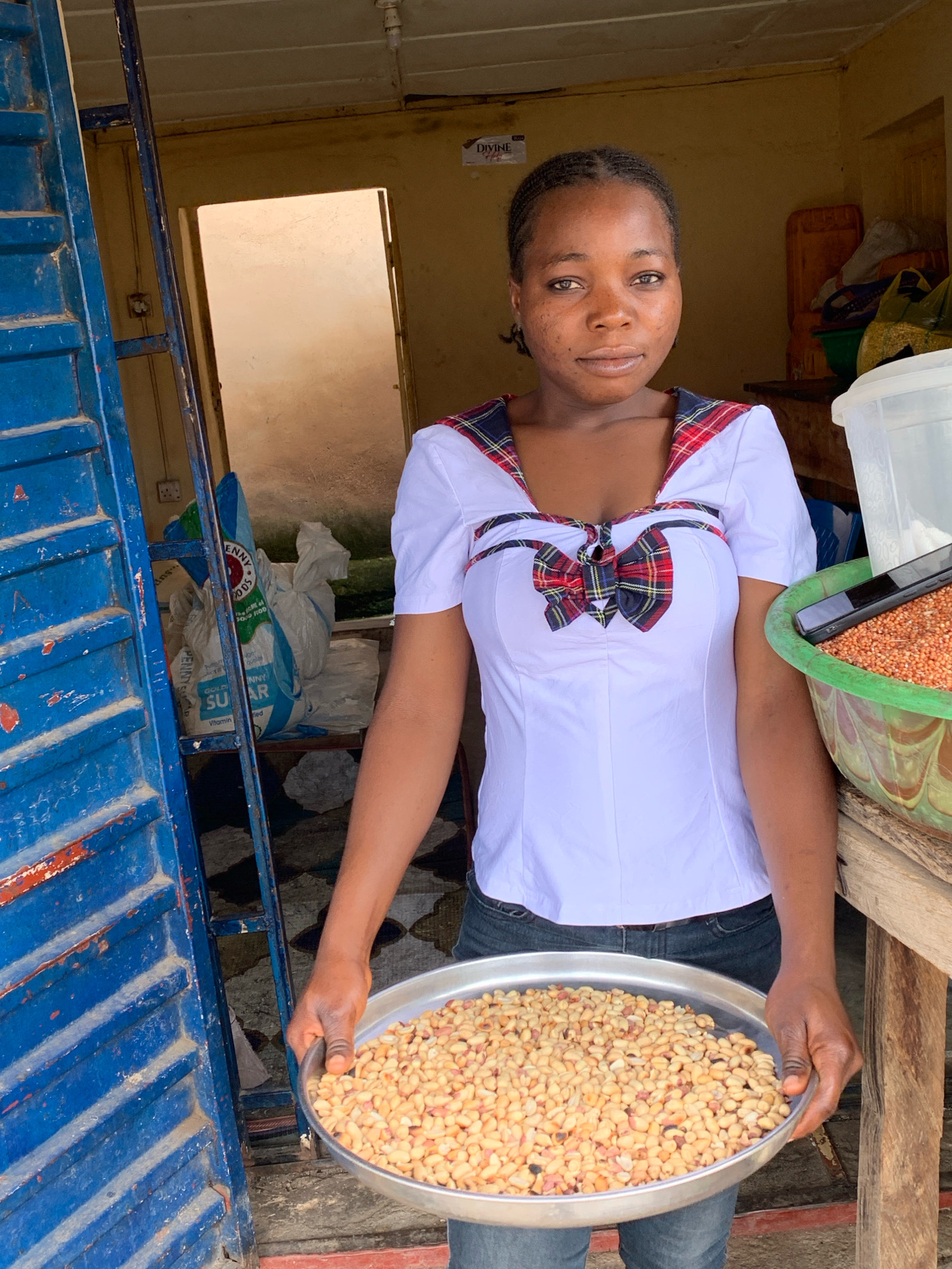 A young Black woman is standing in an outdoor market stall in Jos, Nigeria, holding a large metal tray filled with peanuts. She is wearing a white shirt with a red and black plaid collar and a matching bow tie. The stall appears to be made of wood and metal, with blue corrugated metal on the left side. Bags of sugar are visible behind her, as well as a large green bowl filled with what appears to be grain. A phone is resting on the green bowl. The background suggests an indoor space with a doorway leading to another area. The overall lighting suggests a daytime scene.
