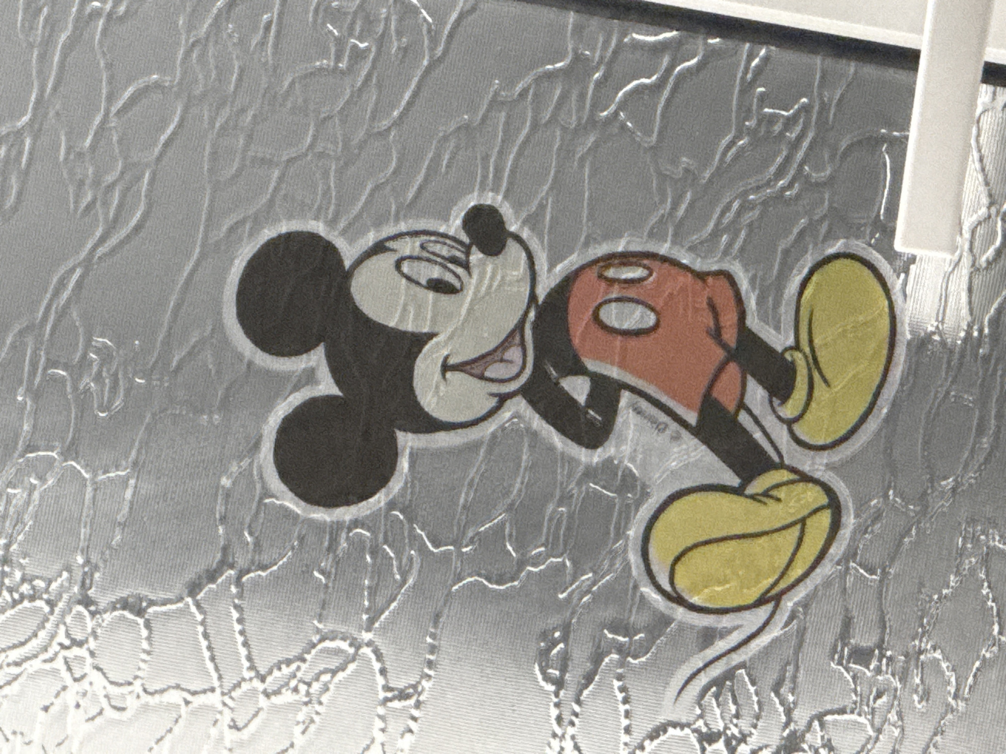 The image shows a depiction of Mickey Mouse against a textured, reflective silver background. Mickey is lying on his side, wearing his iconic red shorts, yellow shoes, and black ears and head. The textured background has a repeating, organic pattern resembling cracked or crinkled material. A small section of white material is visible in the upper right corner of the image. There is no information indicating this image was taken in Shumen, Bulgaria.