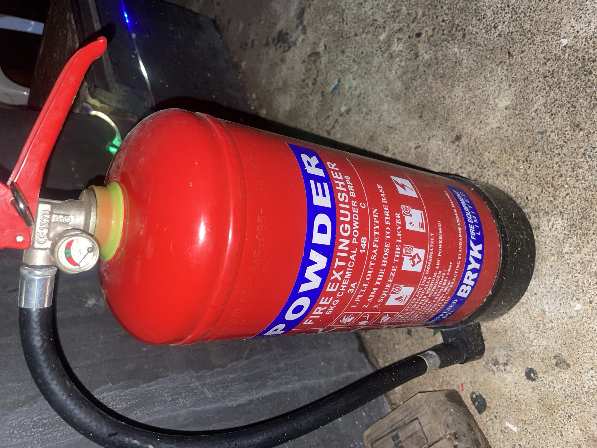 An upright, red, 6kg powder fire extinguisher with a black hose attached to a silver fitting is positioned on a rough, grey, concrete surface. A small, circular pressure gauge with red, green, and yellow indicators is mounted on the top fitting of the extinguisher. The extinguisher has a blue band near the top with white lettering that reads "POWDER FIRE EXTINGUISHER". Below this, smaller text details its chemical composition and usage instructions, including "6KG CHEMICAL POWDER BRP6 3A 14B C", and numbered steps: "1. PULL OUT SAFETYPIN", "2. AIM THE HOSE TO FIRE BASE", and "3. SQUEEZE THE LEVER". Further down, the brand name "BRYK" is printed in white on a blue band. The background shows a dark, textured wall. The image appears to be taken at ground level.