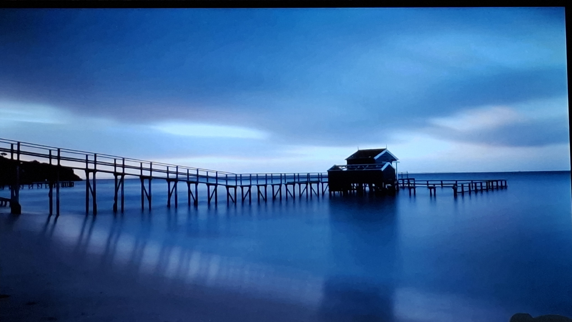 The image presents a serene coastal or lakeside landscape, characterized by various shades of blue. A long, dark wooden pier with handrails extends from the left side of the frame, traversing the water to a small, dark-colored wooden building situated on stilts over the water in the mid-right. This structure appears to be a boathouse or similar utility building. The pier continues for a shorter distance past the building before ending.
The water surface is remarkably smooth and calm, suggesting a long-exposure photograph, creating soft, elongated reflections of the pier supports and the building. The sky above is overcast, featuring soft, streaky clouds in hues of light blue and grey, contributing to a diffused light without direct sunlight. This light quality, combined with the overall blue palette, indicates either dawn, dusk, or an entirely overcast day. No people are visible, and there is no discernible activity taking place, reinforcing the tranquil and contemplative mood of the scene. While the image itself lacks explicit geographical markers, the provided context identifies the location as Saint Petersburg, Russia. There is no visible text within the image.