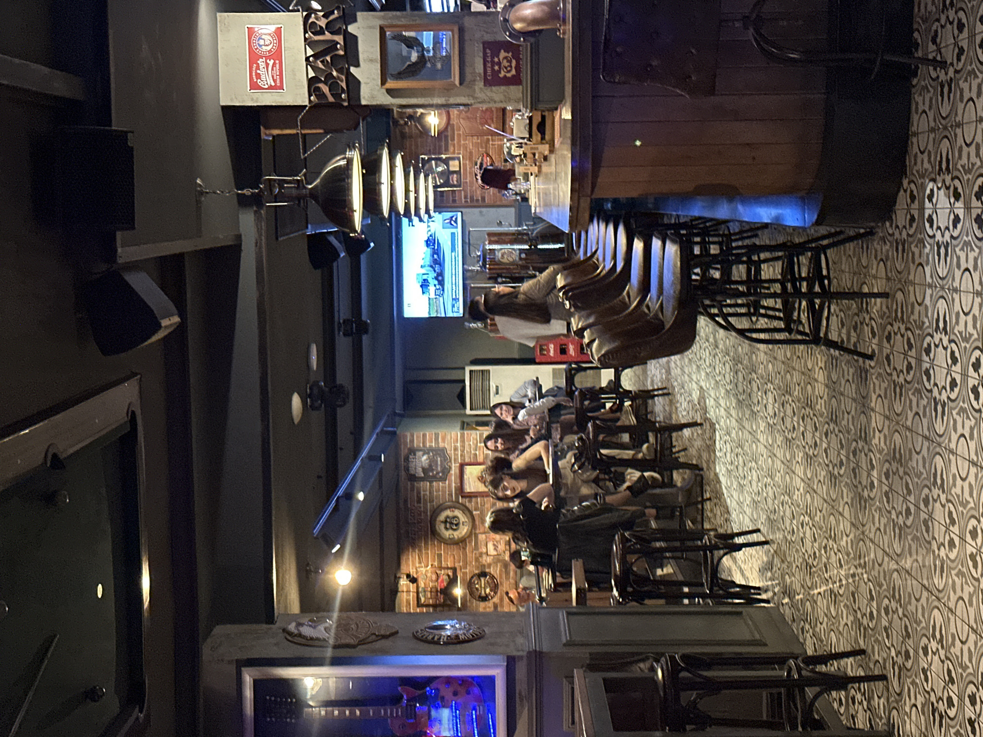The image depicts the interior of a bar with a rock-and-roll theme in Veliko Tarnovo, Bulgaria. The space features a prominent bar counter with several stools, a dance floor, and seating areas. The walls are decorated with various posters and artwork, including a framed electric guitar, and a large screen displays a video of trucks. There are several people seated at tables, engaged in conversation and enjoying drinks. The lighting is dim, with spotlights illuminating the bar and tables. The overall ambiance is casual and relaxed, suggesting a lively evening at the establishment.