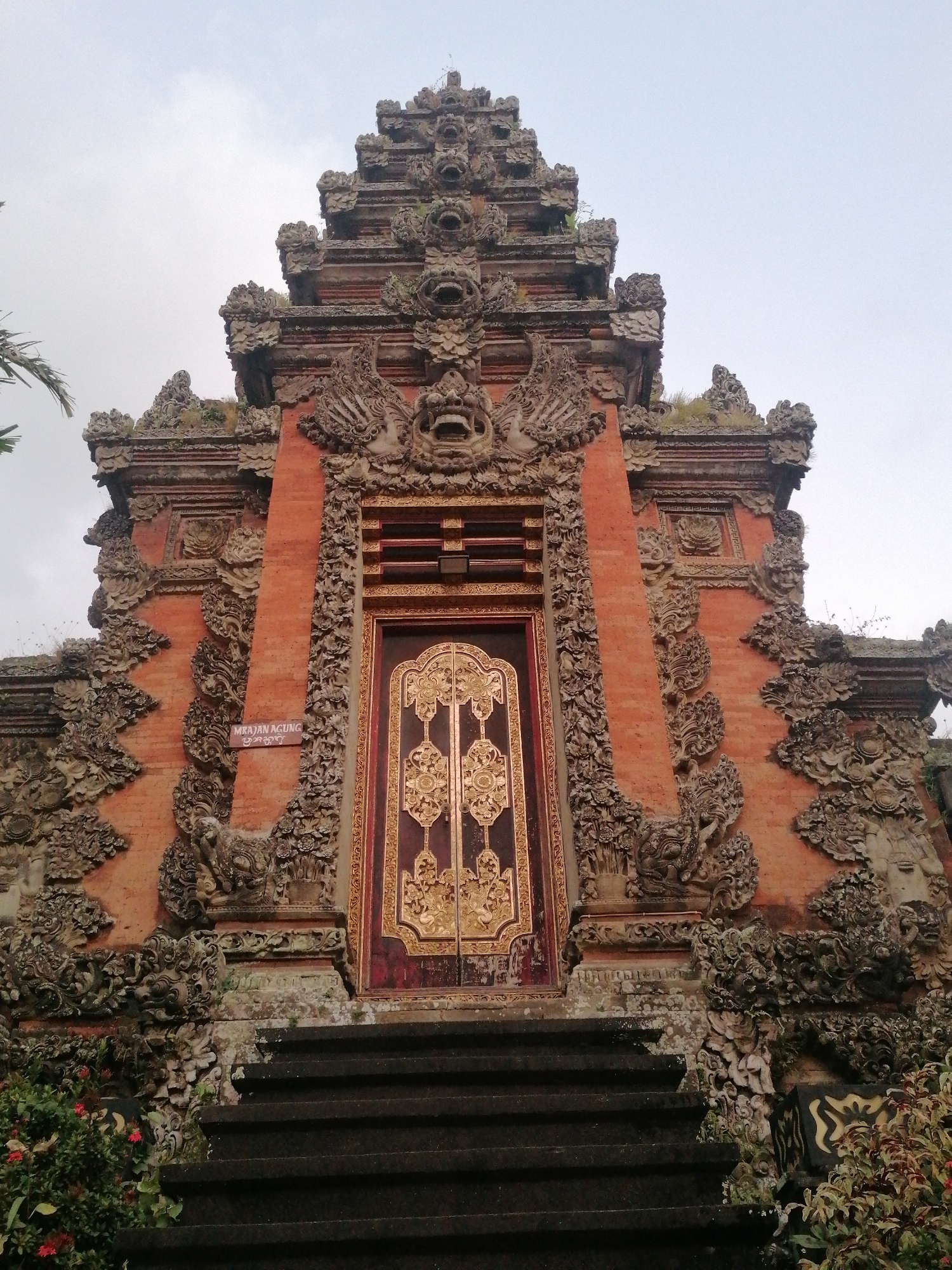 A low-angle, full shot captures the intricately carved facade of a Balinese temple entrance, likely a Candi Bentar or similar structure in Ubud District, Indonesia. The temple is constructed primarily of reddish-orange brick, adorned with detailed stone carvings of mythical creatures, floral motifs, and human-like faces, some of which appear weathered. The central focus is a large, ornate wooden door, painted a deep red and intricately decorated with gold leaf patterns. A sign with text that reads "MRAJAN AGUNG" is affixed to the left side of the entryway. Wide, dark stone steps lead up to the doorway. Lush green foliage with small red flowers spills into the foreground on the lower left and right sides of the frame, partially obscuring the base of the temple structure. The sky above is overcast and pale.