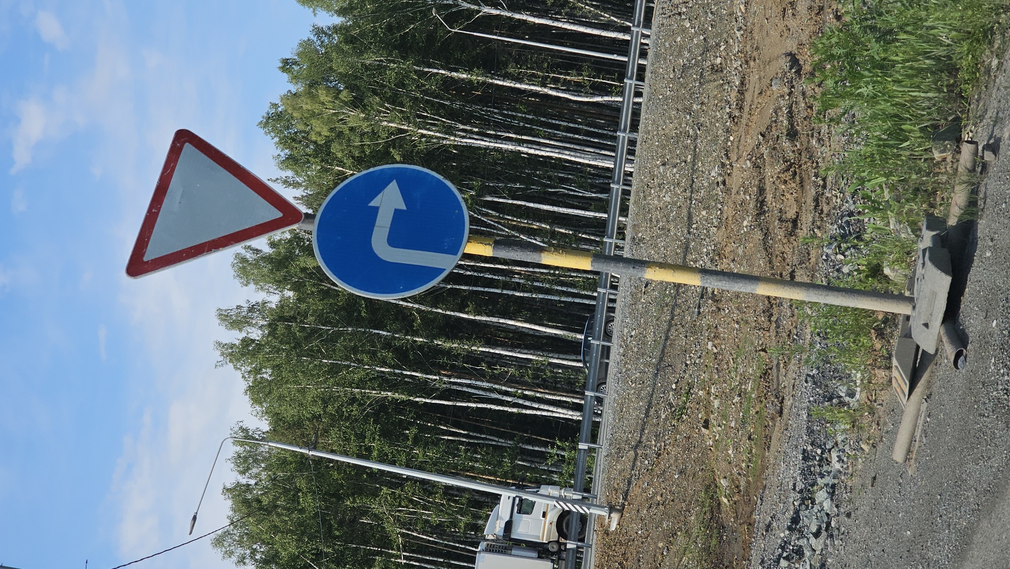 The image shows a roadside scene with traffic signs and a forest in the background. The main subjects are two traffic signs: a triangular sign with a red border and a white interior, and a circular blue sign with a white arrow indicating a mandatory left turn. Both signs are mounted on a metal pole with yellow and gray bands.
The setting appears to be a rural or semi-rural area, likely along a road, with a dense forest of birch trees visible behind a metal guardrail. The ground beside the road is covered in gravel and dirt, with patches of green grass. The sky is blue with some white clouds, suggesting fair weather.
In the foreground, the base of the signpost is visible, anchored in concrete. Further back, a white truck is partially visible on the road, suggesting that this is an active traffic area. A street lamp is also present in the scene.
The image is taken from a slightly low angle, looking upwards towards the signs and the sky. The overall impression is one of a typical road scene in a natural environment. There is no discernible text in the image, other than the symbols on the traffic signs. The time of day appears to be daytime, given the brightness and the clear sky.