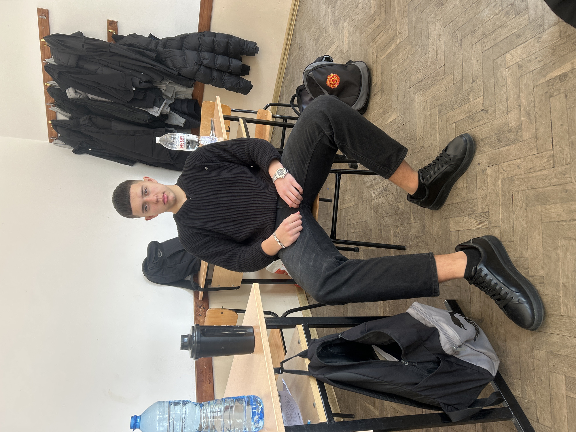 An interior image taken in a classroom in Zlatograd, Bulgaria, shows a young male seated on a chair at a wooden student desk, looking directly at the camera. The male, appearing to be a teenager or young adult, has short, dark hair with a blunt fringe and wears a black ribbed v-neck long-sleeve sweater, dark jeans, and black sneakers with black socks. A silver-toned watch is on his left wrist, and a silver-toned chain bracelet is on his right.
To the immediate left on the desk are a large clear plastic water bottle with a blue cap and a black shaker bottle. A black and grey backpack, featuring a Puma logo, hangs from a hook beneath this desk. Another black backpack, displaying a red Manchester United logo, rests on the wooden floor to the right of the seated individual.
The background features plain white walls, with some faint Cyrillic graffiti visible on the lower left wall. A wooden coat rack mounted on the far wall holds multiple dark-colored winter jackets. A second wooden student desk and chair are partially visible behind the seated male, with another clear plastic water bottle, bearing Cyrillic text including "ГОРНА БАНЯ", positioned on that desk. The floor is covered with worn wooden parquet in a herringbone pattern.