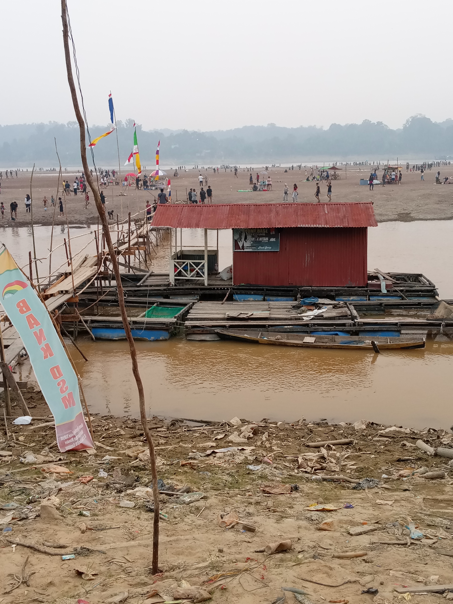 The image depicts a wide, muddy river in Kapuas, Indonesia, with a sandy bank crowded with people. On the river, a red-roofed floating structure serves as a building, possibly a shop or restaurant, with a sign on its side. Several boats are moored around it, some appearing to be on pontoons. A wooden walkway extends from the bank to the floating structure, and a banner with text in an unfamiliar script is visible on the left side of the image. The riverbank is littered with debris, including plastic bags, bottles, and branches. Numerous people are scattered across the sandy area, some walking, others standing or sitting in groups. In the background, a tree-lined horizon suggests a rural or natural landscape. The overall impression is of a lively, informal gathering or market by the river.