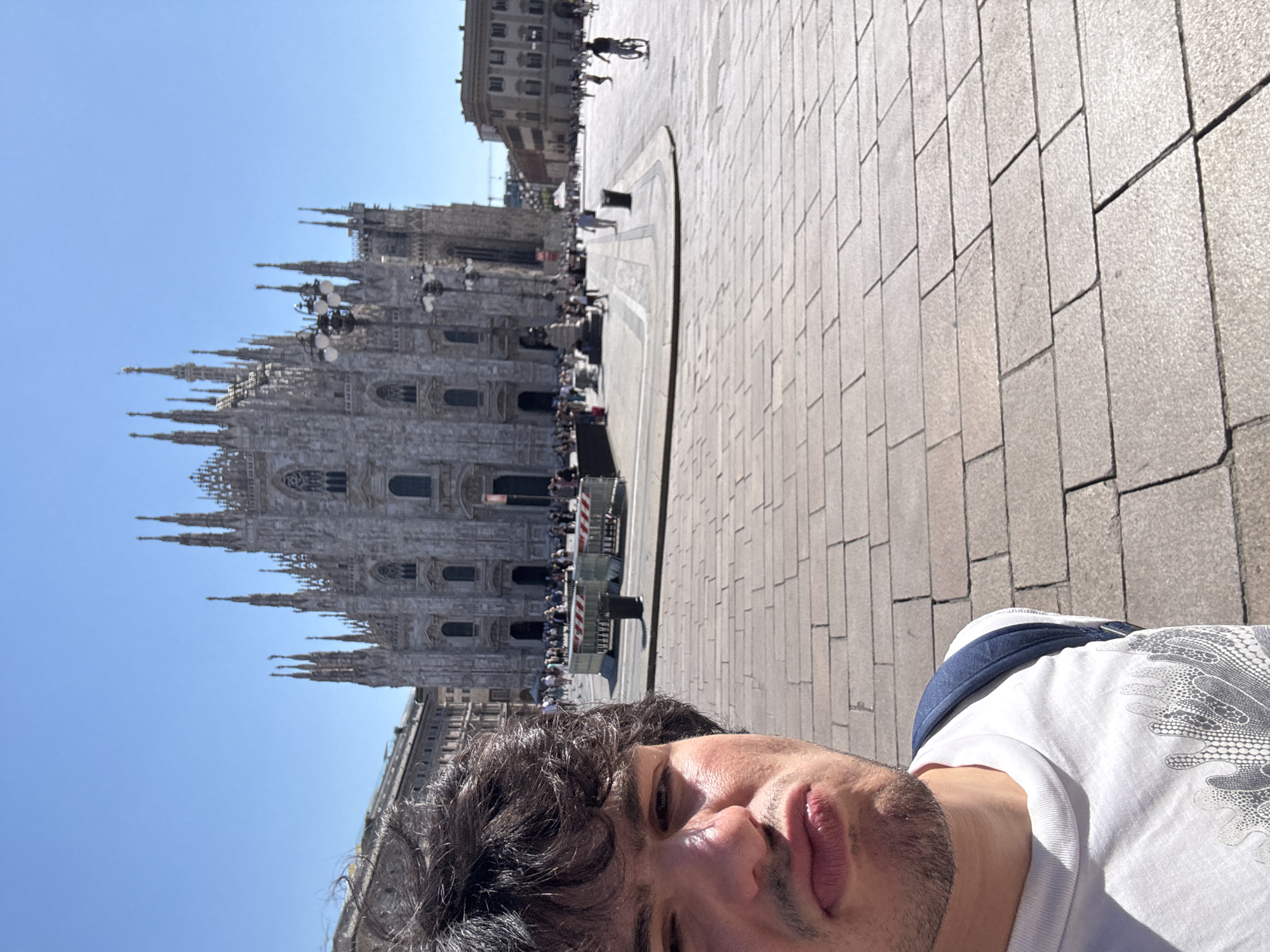 A man is taking a selfie in front of the Duomo of Milan. The man is in the foreground, looking at the camera with a pouty expression and wearing a white t-shirt and a blue backpack. Behind him, the Duomo, a large Gothic cathedral, dominates the background. The plaza in front of the Duomo is paved with stone, and there are several people walking around. The sky is clear and blue.
