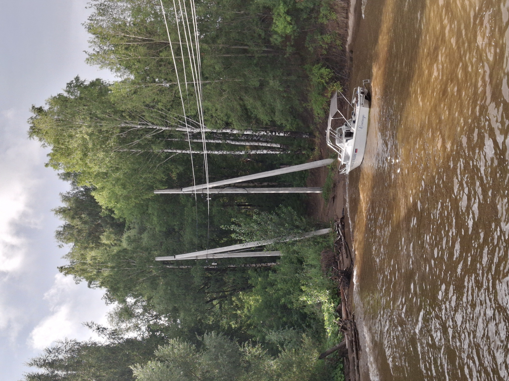 A white motorboat with a gray canopy is docked on the shore of a muddy river, with a lush green forest bordering the bank. Several tall, concrete utility poles stand in the water near the shore, connected by overhead wires. The sky is overcast with scattered clouds. The boat has the name "Courage" written on its stern. The location is the Kama River in Chaikovsky, Russia.