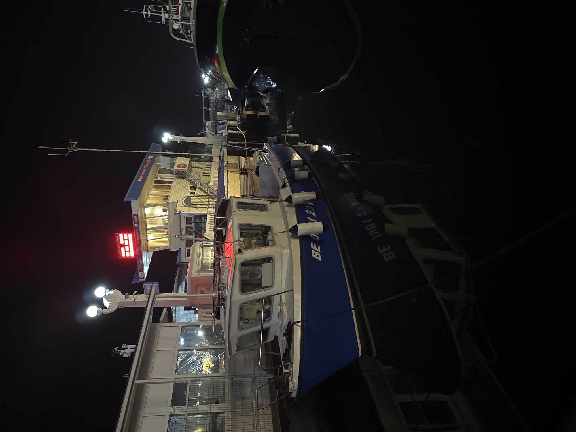 In the city of Sochi, Russia, at night, a small blue and white boat is docked in a harbor. The boat has "BE 0891 27" printed on its side. Another larger boat with a dark hull and a green stripe is partially visible to the right of the smaller boat. In the background, a building with a red illuminated sign that reads "GRAND MARINA" is visible, along with lights from other structures and streetlights. The scene is illuminated by artificial lights, creating reflections on the dark water.