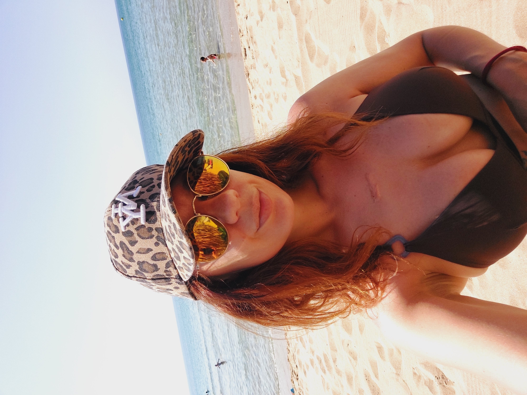 A woman with auburn hair is taking a selfie on a sandy beach in Sozopol, Bulgaria. She is wearing a leopard-print baseball cap, mirrored sunglasses, and a dark bikini top. The sea is visible in the background, with a few people swimming. The sun is shining, casting a warm glow on the scene.