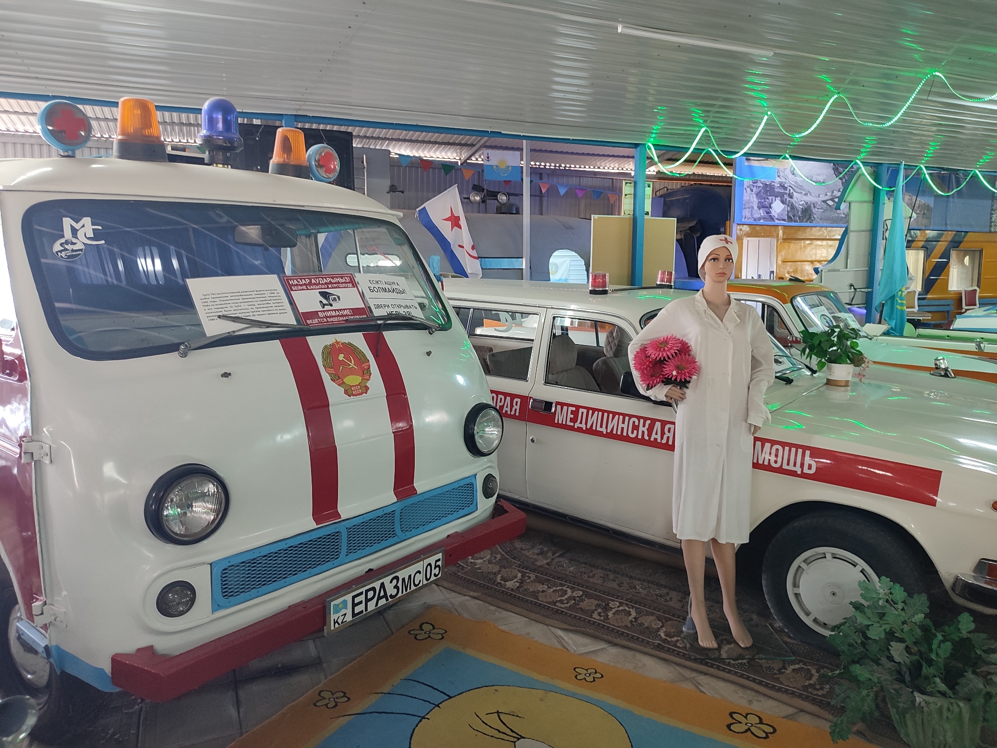 In Qonaev, Kazakhstan, within a museum or exhibition space, two vintage ambulances are displayed side-by-side. The ambulance in the foreground is white with a prominent red stripe running down its center, featuring a Soviet-era emblem. It has flashing lights on its roof, including red cross symbols, amber, and blue lights. The second ambulance, partially obscured, also appears white with red markings and the Cyrillic text "МЕДИЦИНСКАЯ ПОМОЩЬ" (Medical Assistance) visible on its side. A mannequin dressed as a nurse, wearing a white uniform and cap and holding a bouquet of pink flowers, stands beside the second ambulance. The background reveals a tent-like structure with colorful pennant banners and a large screen displaying an image. Green LED lights are strung across the ceiling. The floor is covered by a patterned rug and a brightly colored mat with cartoon imagery.