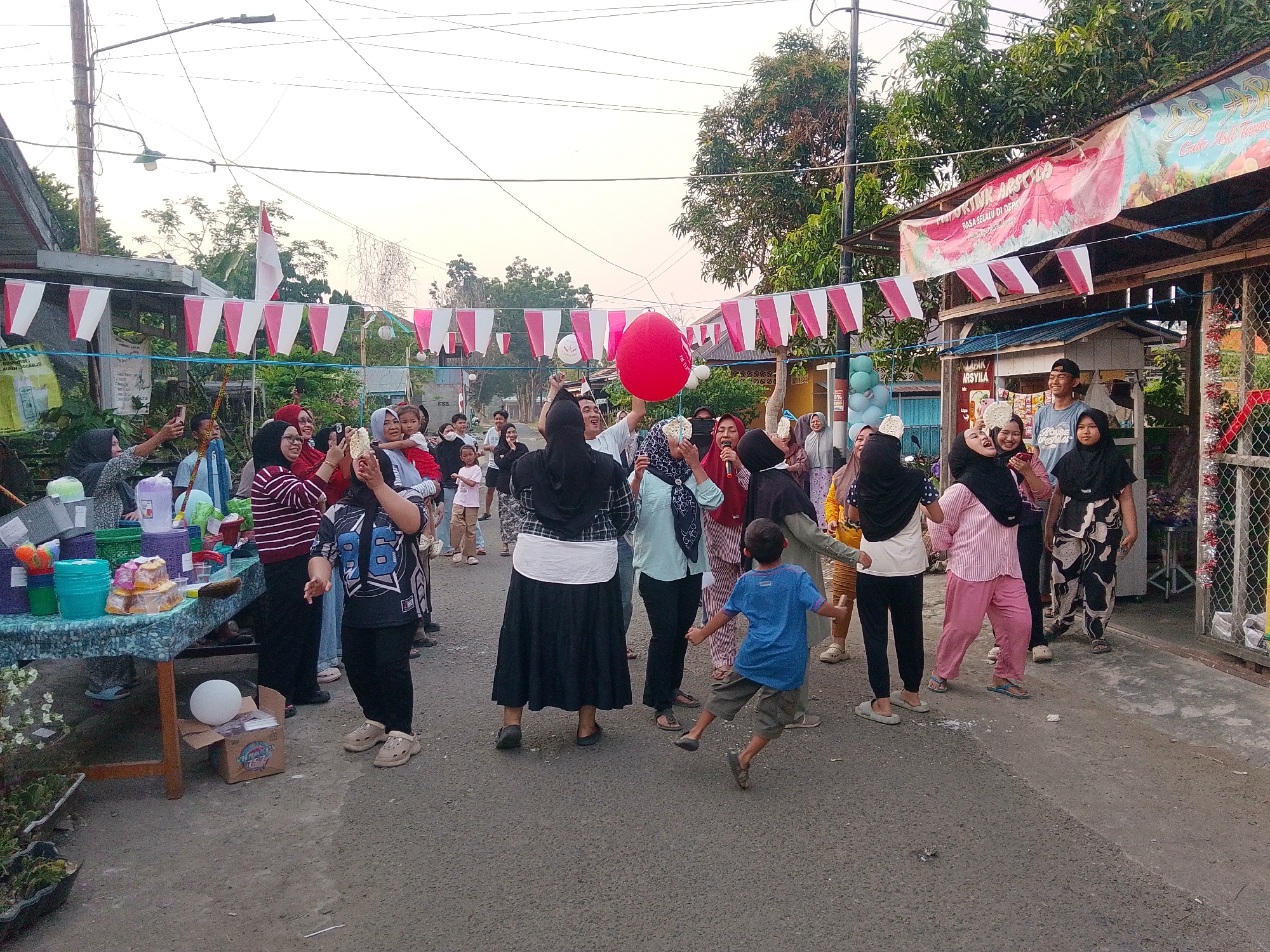 In Barabai, Indonesia, a group of people are gathered in a street, likely for a local celebration or event, as indicated by the festive decorations. Red and white pennant flags are strung across the street, and a large red balloon is being held up by several individuals. The sky is overcast, suggesting it is late afternoon or early evening.
The scene is populated by a diverse mix of individuals, including children and adults, many of whom are wearing casual clothing, with some women in hijabs. Several people are holding up their phones, presumably to capture the moment, and some are interacting closely, laughing and talking.
To the left of the frame, a table is set up with various items, including containers, what appear to be snacks or small food items stacked on plates, and a white ball in a cardboard box. Further to the right, a shop or stall is visible, adorned with more festive decorations, including tinsel and balloons. The ground is paved with asphalt. The overall atmosphere appears to be one of joyful community engagement.