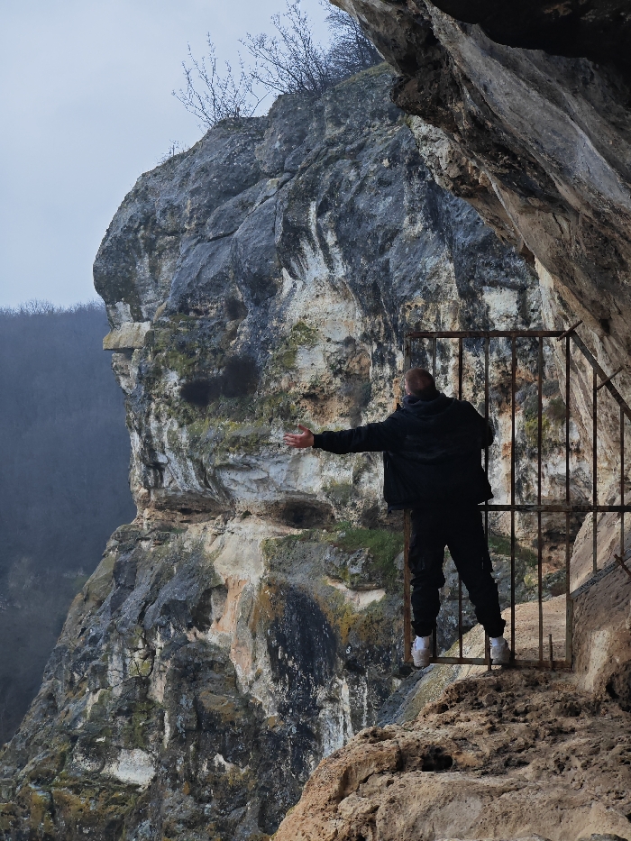 An adult male stands with his back to the camera at the entrance of a rock shelter, part of the Osmar Rock Monastery complex in Shumen, Bulgaria. He is positioned on a rugged, uneven rock ledge within the cave opening, facing outwards towards a valley. The man wears a black hooded jacket, black cargo pants, and white athletic shoes. His arms are extended horizontally to his sides, palms open. Behind him, a dark, weathered metal gate consisting of vertical bars is affixed to the natural rock face, securing the opening. The immediate environment is composed of light-colored limestone rock, heavily weathered with visible stratification and patches of dark moss or lichen. Above the man, the cliff face extends upwards, topped with sparse, bare tree branches against an overcast, light grey sky. The distant background reveals a forested valley and hills, partially obscured by atmospheric haze.