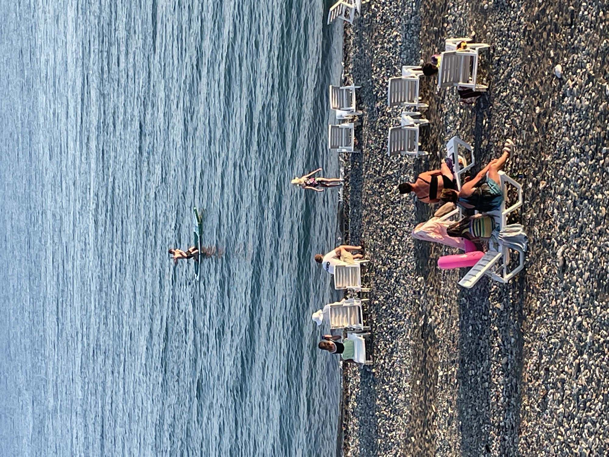 The image depicts a sunny day at a pebble beach in Gagra, Abkhazia. The sea, a vibrant blue, is calm with gentle waves. In the water, a person is stand-up paddleboarding. Closer to the shore, several beach chairs are arranged on the pebbled beach. Some chairs are occupied by people relaxing, while others are empty. A few individuals are visible wading in the shallow water at the edge of the sea. Beach accessories such as towels and inflatable toys are scattered among the chairs, suggesting a leisurely atmosphere. The sunlight casts long shadows across the beach, indicating it is likely late afternoon or early morning.