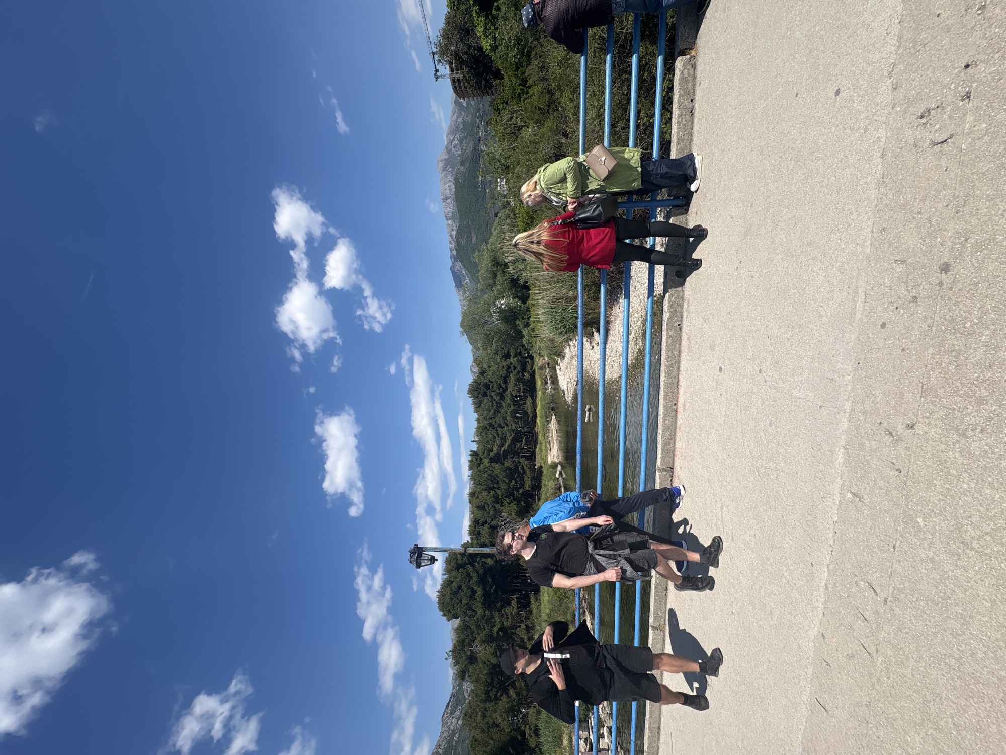 The image shows a sunny outdoor scene from Bar, Montenegro. Several people are standing on a bridge with blue metal railings. To the right are two women, one in a green jacket and another in a red jacket, both leaning against the railing. Further along the bridge, closer to the camera, are three men, dressed in casual attire. The backdrop features a landscape of trees and mountains under a blue sky dotted with fluffy white clouds. A river or stream can be seen running under the bridge. The pavement of the bridge is grey, and the shadows of the people are cast onto it. There is a street light pole, possibly a lamp post in line with the men standing on the bridge.