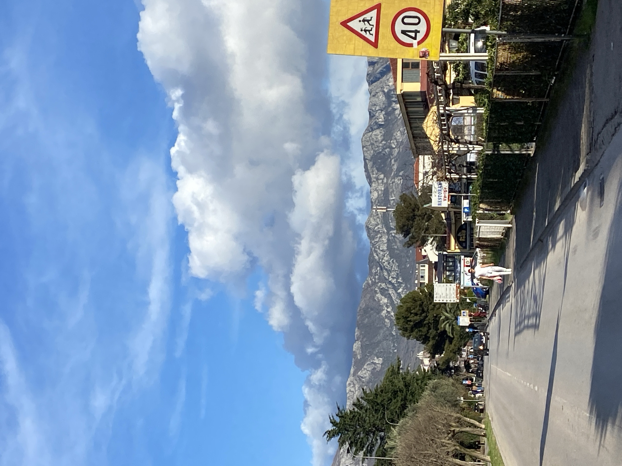 The image depicts a street scene in Budva, Montenegro, under a partly cloudy sky. A two-lane asphalt road with a dashed white centerline extends into the distance from the foreground. On the left, a sidewalk is bordered by a row of mature deciduous trees with bare branches and some evergreen shrubs. Further down the road, several indistinct pedestrians and a dark-colored car are visible.
On the right side of the road, a paved sidewalk runs parallel to a metal fence densely covered with green foliage. A male individual, dressed in a white t-shirt and white pants, walks away from the camera on this sidewalk. Immediately adjacent to the sidewalk on the right stands a prominent yellow signpost. This post displays two signs: a red triangular warning sign with black silhouettes of two children, and a circular speed limit sign indicating '40'.
Behind the fence and signs on the right, several low-rise buildings with light-colored facades and orange/yellow tiled roofs are present. One building features a sign reading "RESTORAN Vertigo". In the background, a formidable range of rugged, grey mountains dominates the horizon, partially enveloped by large white and grey cumulus clouds. The sky above is clear blue with scattered clouds. The scene is illuminated by direct sunlight, casting elongated shadows across the road, suggesting a morning or late afternoon timeframe.
