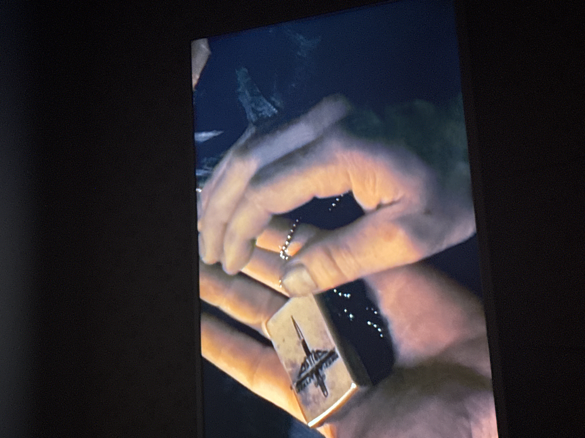 A close-up shot displays a person's hands holding a Zippo lighter and a dog tag chain. The lighter has a dark design etched on a lighter-colored surface. The hands are positioned against a dark, blurry background. No identifiable location or individuals can be determined from the image.