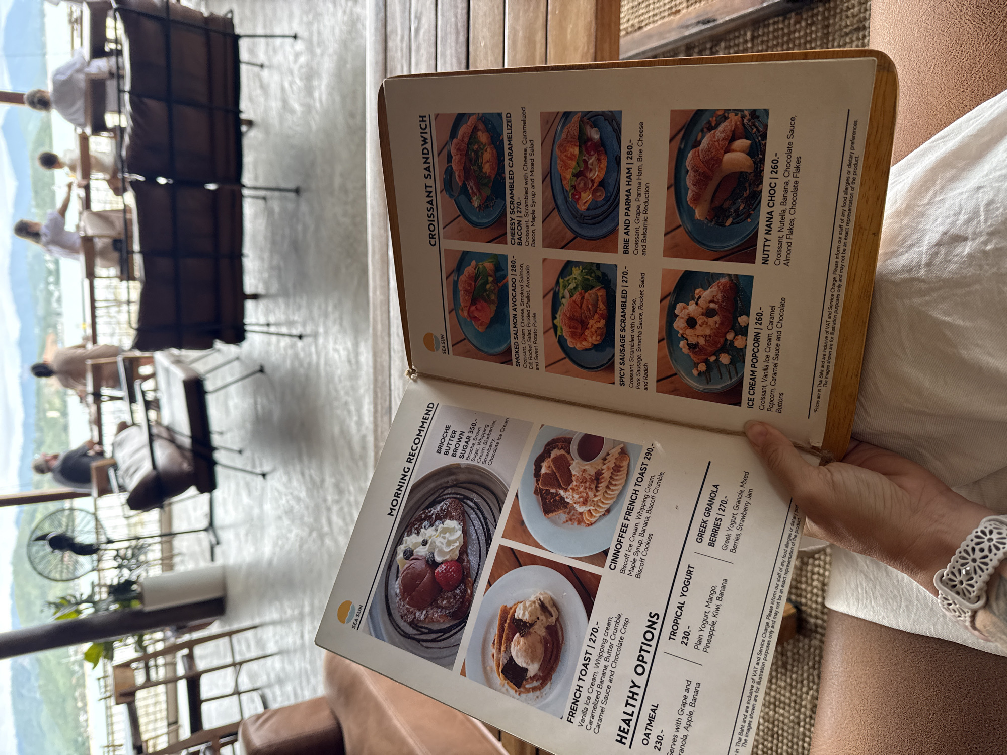 The image shows a person's hands holding an open menu from "SEA SUN" restaurant, which is located in Ko Samui District, Thailand. The menu displays breakfast and lunch options, including "MORNING RECOMMEND" items like Brioche French Toast, "HEALTHY OPTIONS" such as Oatmeal and Tropical Yogurt, and "CROISSANT SANDWICH" varieties. The background reveals an indoor dining area with patrons seated at tables and a view of the ocean and lush green hills, suggesting a resort setting. The overall ambiance is relaxed and casual.