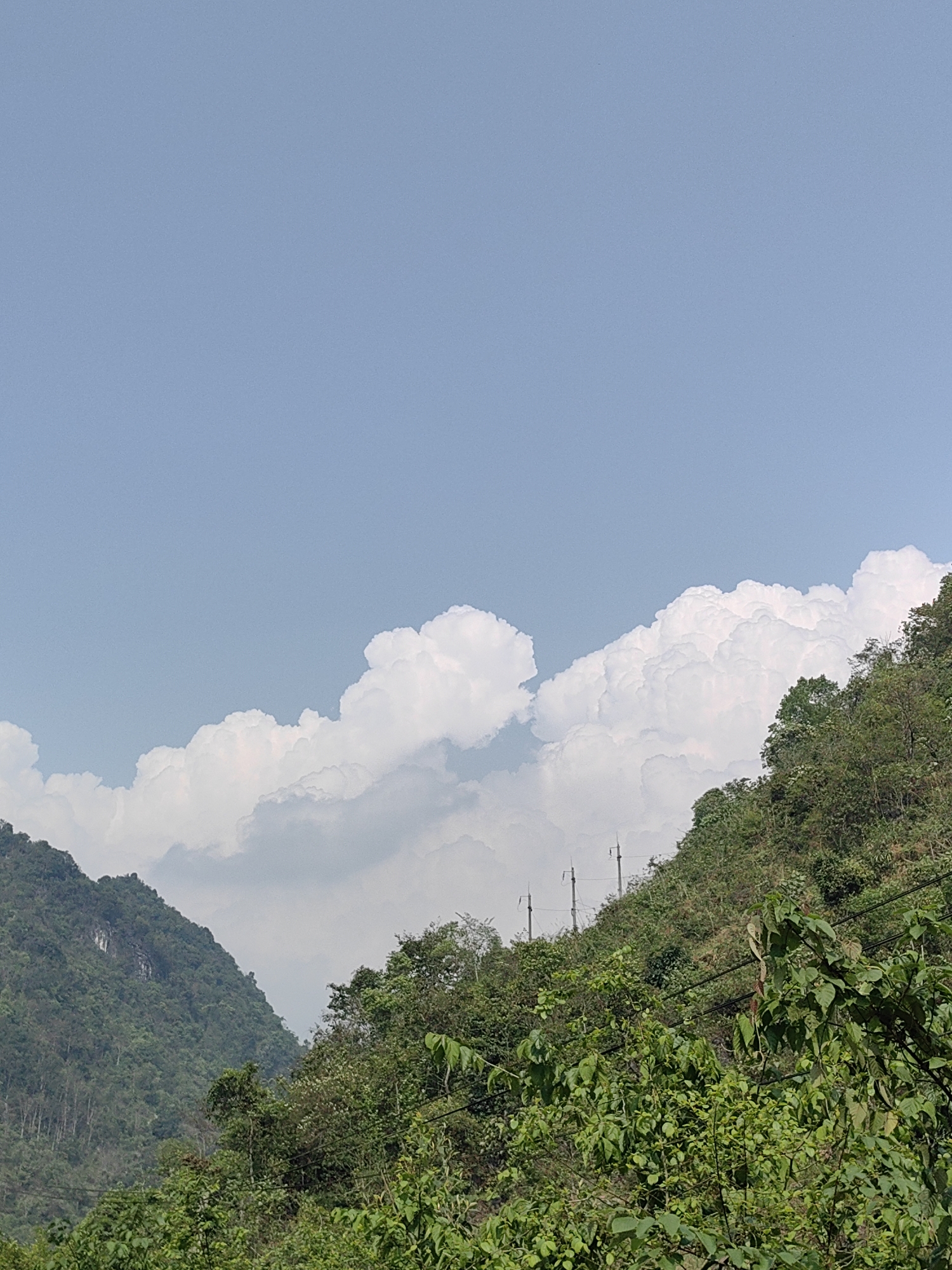 The image depicts a landscape in Xã Hà Quảng, Vietnam. A lush, green mountain rises on the right side of the frame, densely covered with trees and vegetation. Power lines are visible running along the mountainside. To the left, another mountain, less prominent, provides a contrasting backdrop. Above the mountains, the sky is a clear, pale blue, partially filled with large, white, fluffy cumulus clouds. The overall scene conveys a sense of natural beauty with subtle human influences.