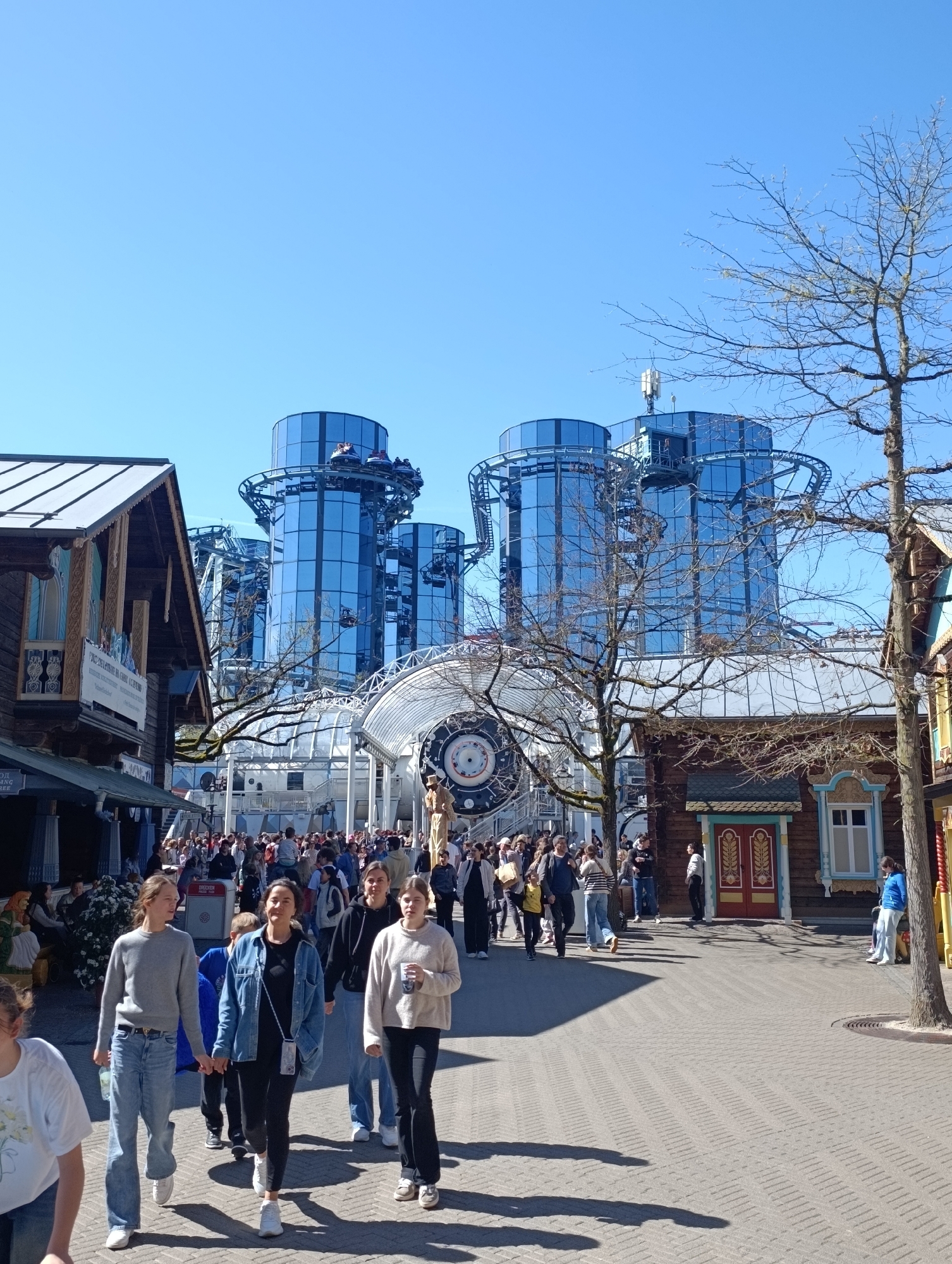 The image shows a bustling scene at Europa-Park, a theme park in Rust, Germany. A large crowd of people is walking along a brick-paved pathway lined with buildings in a rustic style. Dominating the background are tall, cylindrical, glass-clad towers connected by a roller coaster track. There is a large clock-like structure visible behind the crowd. A bare tree is standing to the right. The sky is clear and blue.