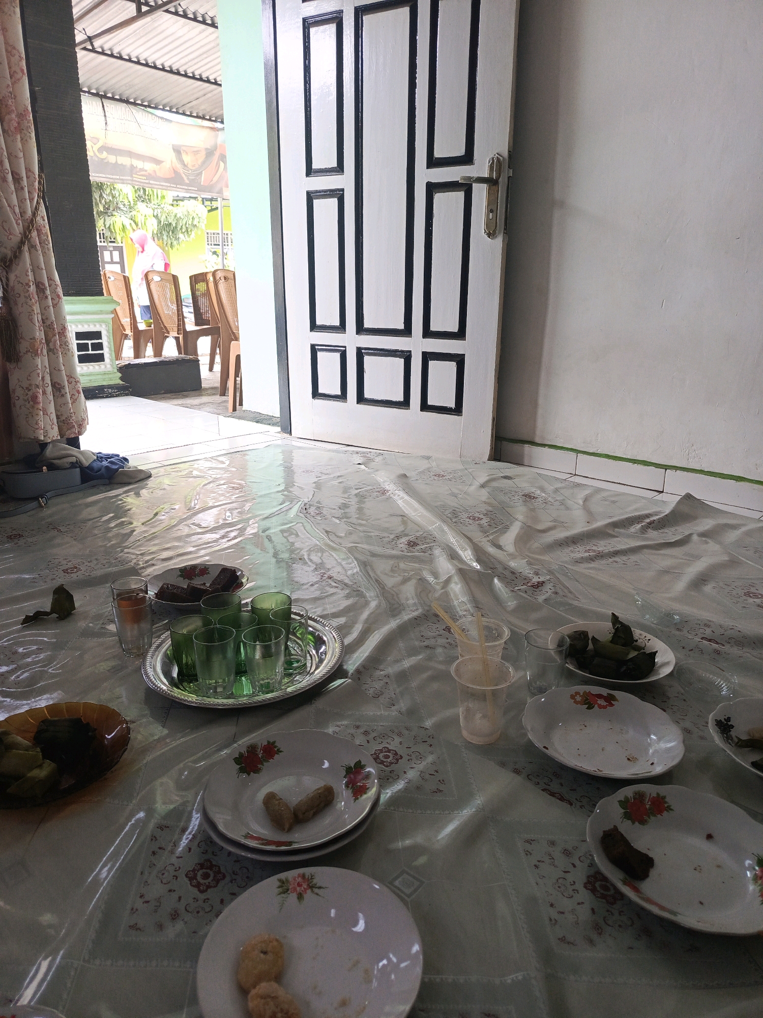 The image depicts a low-angle shot of a room in Lubuklinggau, Indonesia, where food and drinks are laid out on a plastic sheet covering the floor. In the foreground, several plates are visible, some with remnants of food, indicating a meal has recently concluded or is in progress. There are also glasses, some containing liquid, and plastic cups with straws. A silver tray holds several green glasses. To the left, a brown dish contains what appear to be wrapped food items. In the background, a white door with black trim and a brass doorknob is a prominent feature. Beyond the door, an outdoor area is visible with chairs and trees. A patterned curtain hangs on the far left. The lighting suggests daytime, with sunlight streaming in from the open doorway. The overall scene conveys a sense of informal gathering or mealtime within a residential setting.