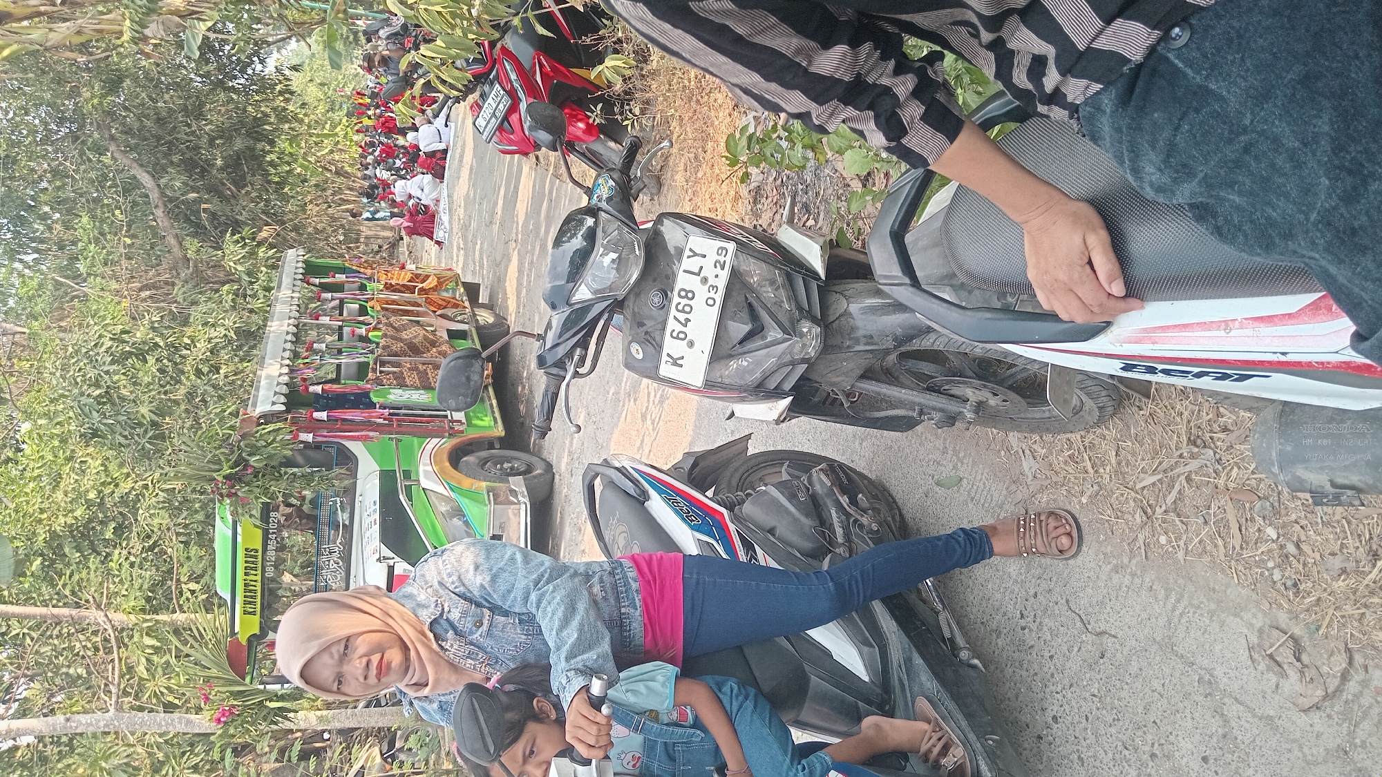 In Kedungwaru, Indonesia, on a sunny day, a woman wearing a hijab and a denim jacket is on a motorcycle. She is accompanied by a young girl in overalls. The background features a green and white bus decorated with flowers and banners, and another motorcycle. In the distance, a crowd of people are gathered, some seated on the ground. The scene suggests a community event or gathering in a rural or semi-urban setting.