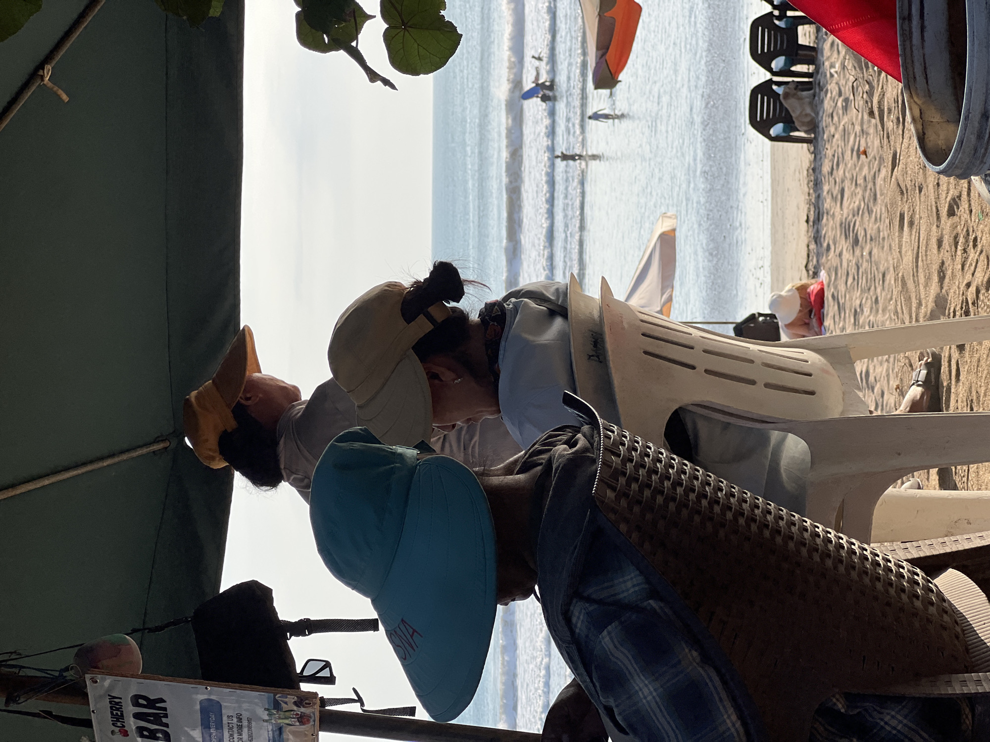 In Kuta, Indonesia, three individuals are seated at tables under a large green umbrella on a beach. In the foreground, a person wearing a plaid shirt and a teal visor faces away from the camera. To their right, another individual in a light-colored shirt and a tan hat is also seated, looking out towards the ocean. Further back, a third person, also wearing a hat, is visible. The ocean stretches out in the background, with gentle waves and several people in the water, some of whom appear to be surfing. Beach chairs and umbrellas are scattered on the sand, and a dog rests near two empty black chairs. A sign that reads "CHERRY BAR" is attached to a pole near the umbrella. The scene suggests a relaxed atmosphere on a sunny day at the beach.