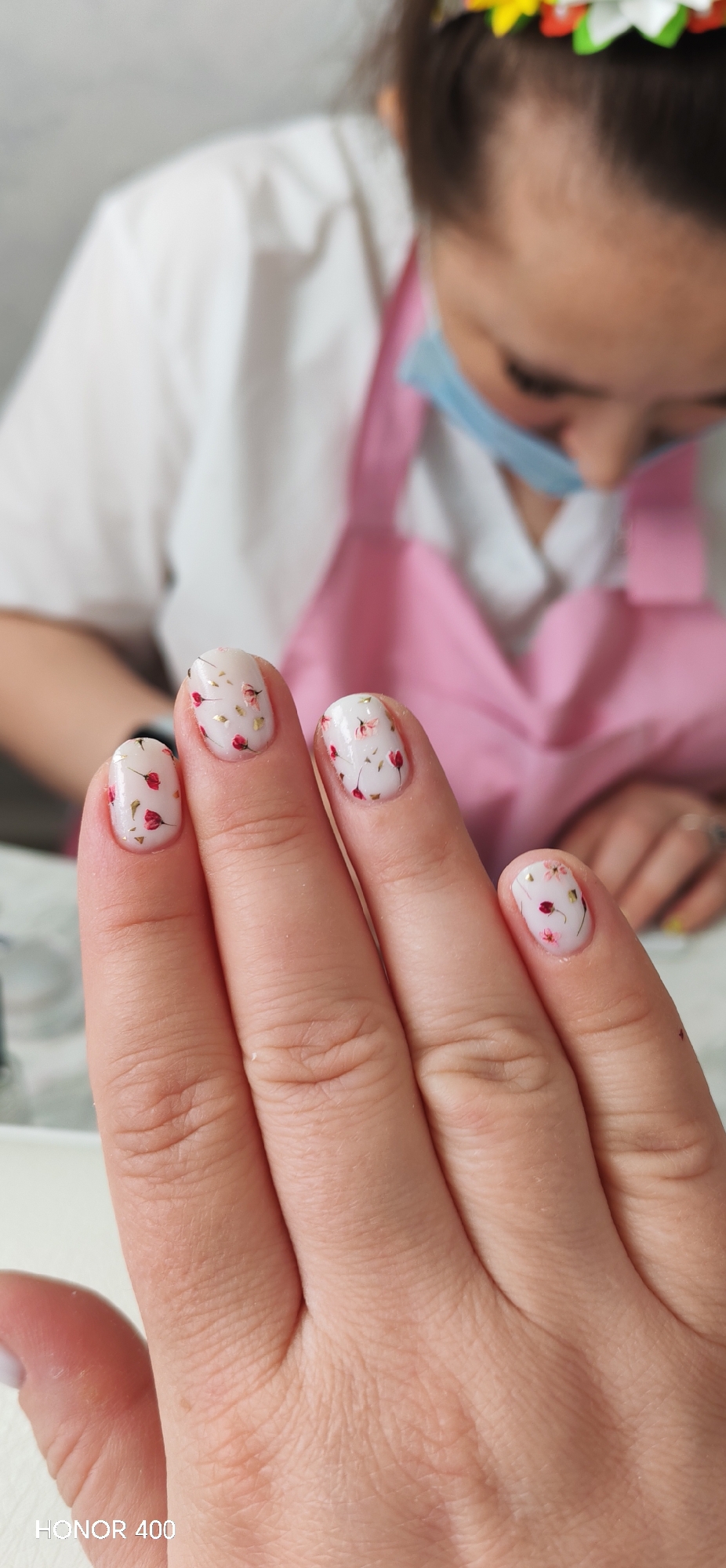 A close-up photograph shows a woman's hand with decorated fingernails, with a manicurist wearing a mask and a pink apron blurred in the background. The fingernails are painted white with delicate red flower and gold leaf designs. The skin on the hand is fair and shows natural skin texture. The background includes a blurred white surface with cosmetic items and tools, suggesting a salon setting. The manicurist's face is partially visible, with her eyes focused downwards. The "HONOR 400" watermark is visible in the lower-left corner. There is no information in the image that can confirm its location in Noviy, Russia.