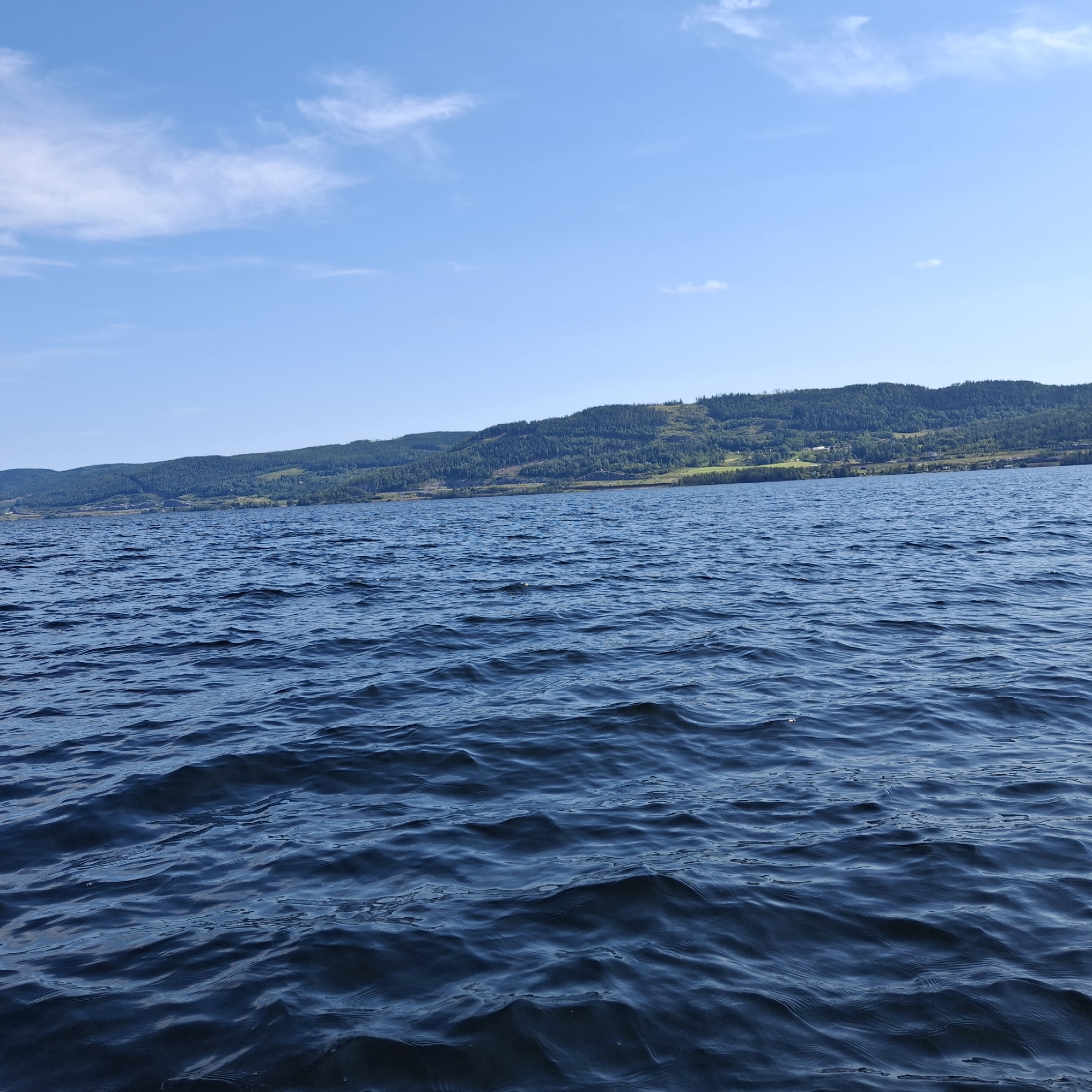 The image shows a wide, deep body of dark blue water, likely a lake or a fjord, with visible ripples and small waves. The water takes up the lower two-thirds of the frame. In the distance, a gently sloping, tree-covered hillside rises from the opposite shore, creating a natural horizon. The hillside is a mosaic of dark green trees, with patches of lighter green indicating clearings or vegetation variations. Some structures, possibly buildings or farms, are visible nestled into the landscape on the hillside. The sky above is a clear, bright blue with a few wispy white clouds scattered across it, indicating fair weather. The scene is a natural landscape, likely captured from a boat or elevated vantage point on the water, depicting the serene environment of Minnesund, Norway, known for its location on the Eidsvoll municipality on the northern end of Norway's largest lake, Mjøsa.