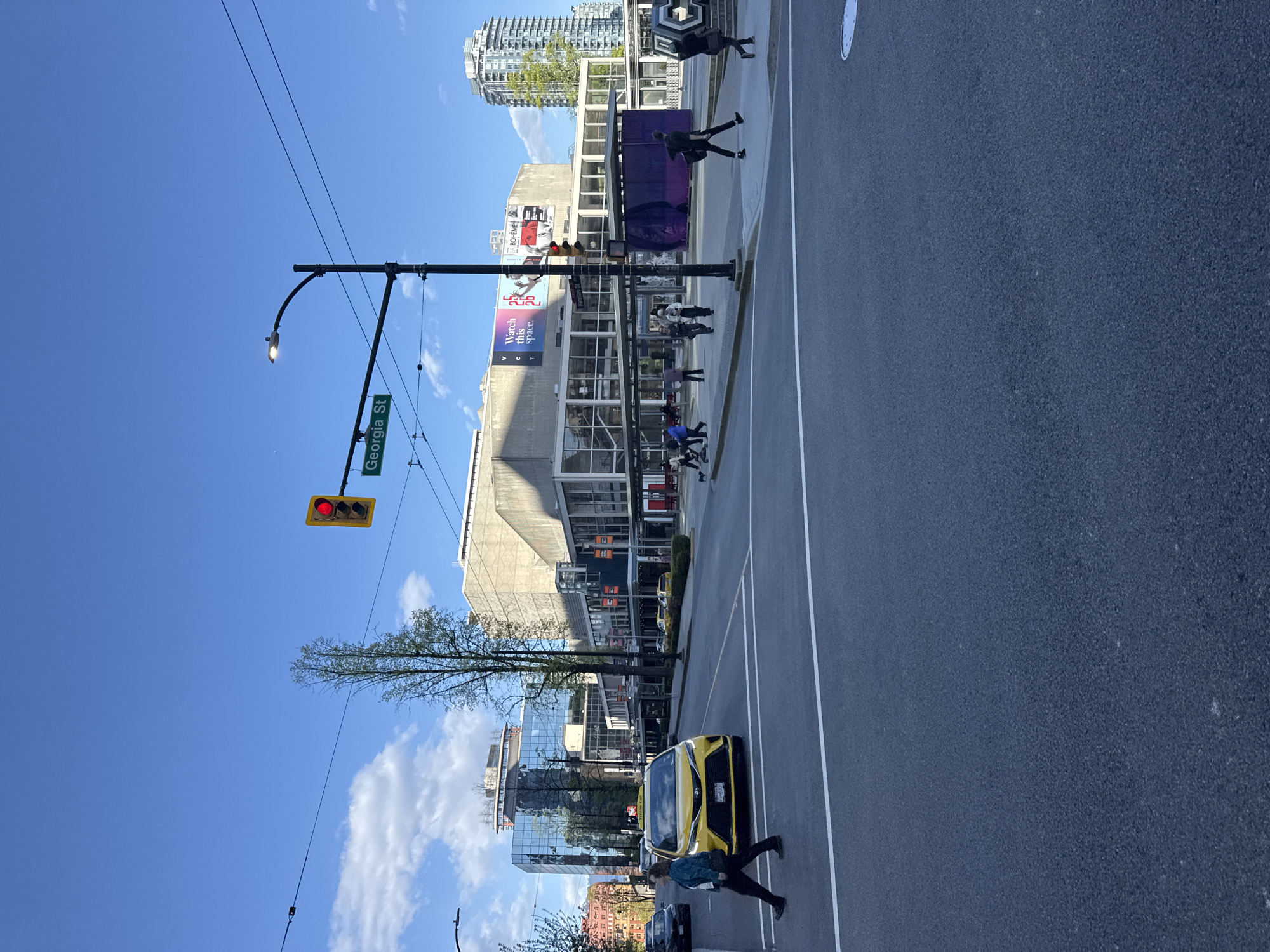The image shows a street scene in Vancouver, Canada, specifically at the intersection of Georgia Street. A red traffic light is visible hanging from a pole with a street sign indicating "Georgia St." People are walking on the sidewalk and crossing the street. A yellow taxi is positioned on the road. Tall buildings dominate the background, with a clear blue sky above.