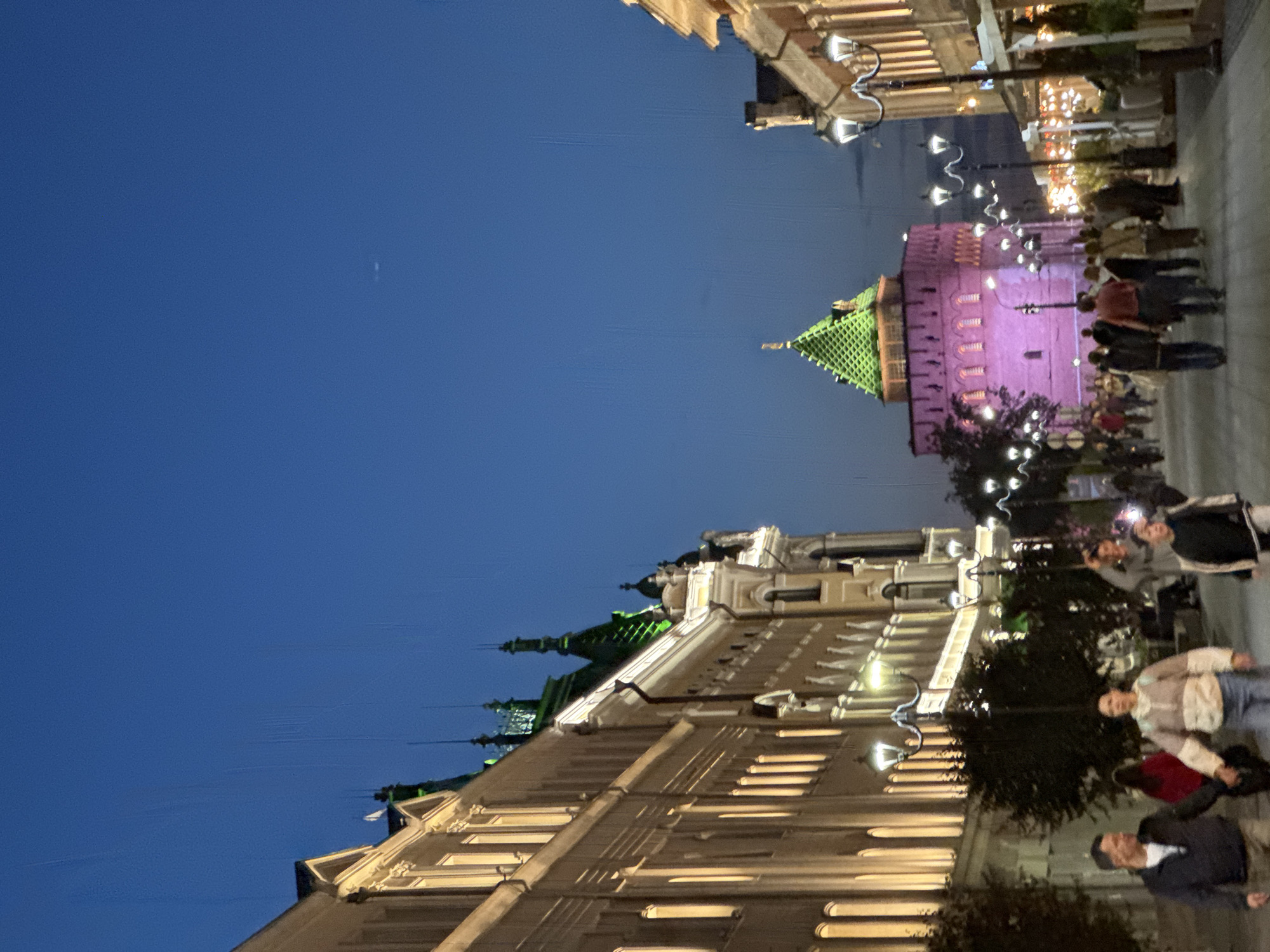 The image depicts a nighttime street scene in Nizhny Novgorod, Russia, likely in the historic city center, possibly near the Kremlin. A pink tower with a green, conical roof is illuminated in the background, a prominent landmark. The street is lined with multi-story buildings on either side. The building on the left is a light-colored structure with numerous windows, some illuminated from within. Ornate architectural details and decorative lighting adorn its facade. The building on the right is also illuminated, with outdoor seating and dining areas visible, suggesting it houses restaurants or cafes. Several streetlights with ornate designs illuminate the pedestrian walkway.
People are walking along the street, some in pairs, holding hands. They are dressed in casual, modern clothing. The sky is a deep blue, indicative of twilight or night. The overall atmosphere is lively and urban, with the architecture and lighting contributing to a festive and inviting ambiance.