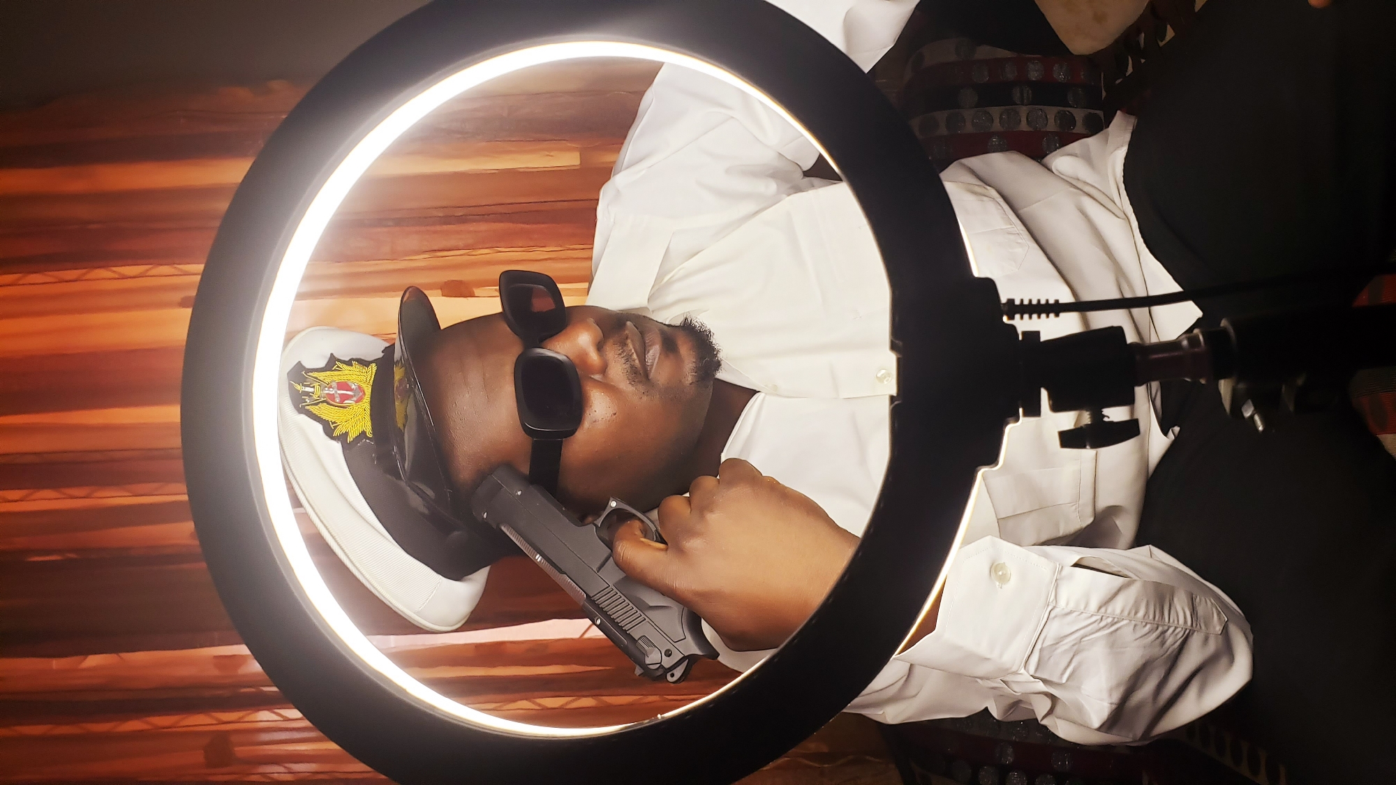A man is centrally positioned within a bright ring light, posing for a photograph indoors. The image is rotated 90 degrees clockwise. The man has dark skin and a short beard, wearing large, dark rectangular sunglasses that obscure his eyes. He is dressed in a white, long-sleeved button-up shirt and dark trousers.
On his head, he wears a dark cap featuring a prominent gold and red emblem, suggestive of a military, police, or official uniform. In his right hand, he holds a black handgun, with his index finger placed on the trigger. The man's expression is serious or neutral, partially hidden by the sunglasses.
The ring light provides strong, even illumination, creating a striking portrait effect. The background consists of horizontal wooden planks or blinds with warm, brownish hues. The setting appears to be an indoor environment in Sabon Tasha, Nigeria. The photograph emphasizes a stylized and intense mood. No visible text can be distinctly read on the cap's emblem or the handgun.